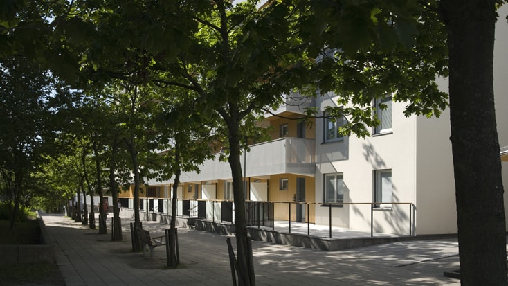 Modern apartment building lined with trees on a sunlit street, featuring balconies and walkways.