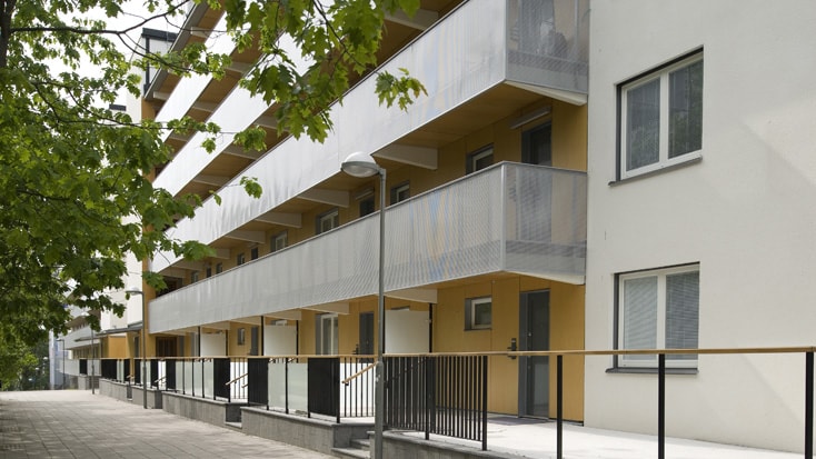 Modern apartment building exterior with yellow facade and glass balconies, surrounded by trees.