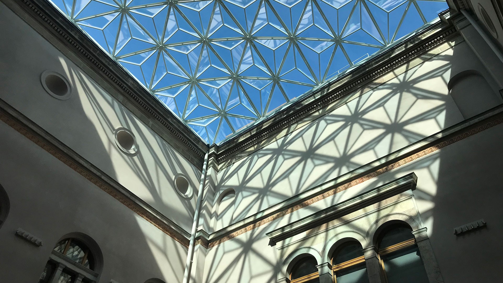 Modern glass dome with geometric patterns casting shadows on historic building's interior walls.