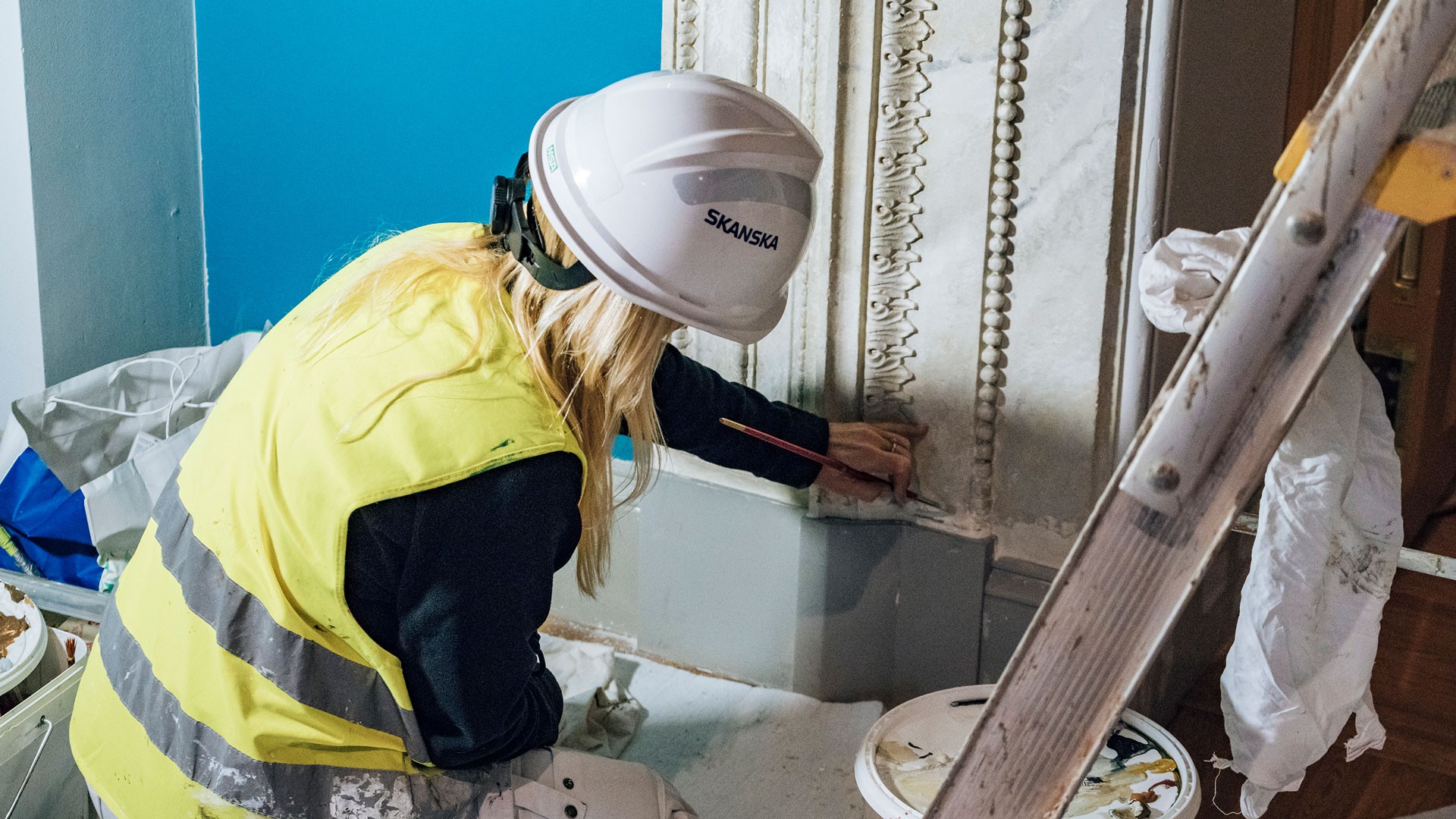 A construction worker in a hard hat and safety vest carefully paints ornate architectural details.