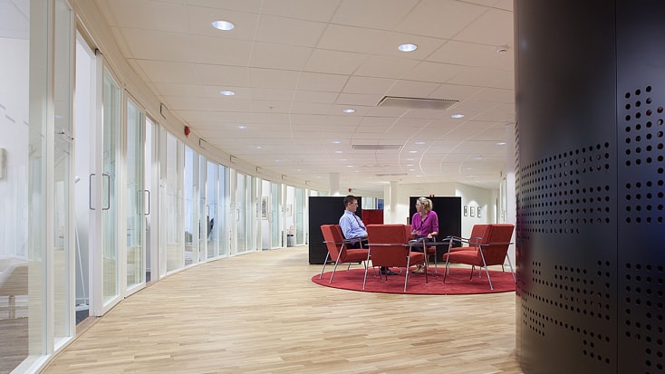 Modern office space with curved glass walls, wooden flooring, and two people seated on red chairs.