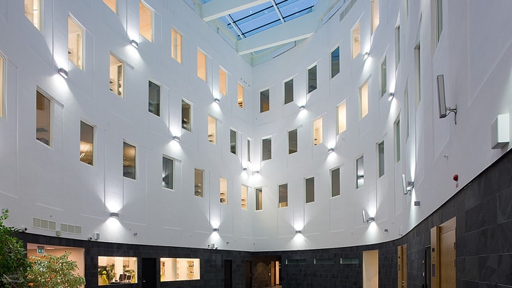 Modern atrium with tall white walls, numerous windows, and skylight, illuminated by wall lights.