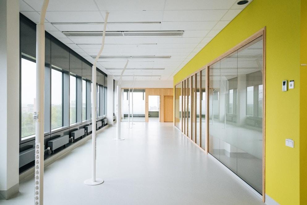 Moderne Bürogang mit großen Fenstern, gelber Akzentwand und deckenmontierten Stangen.