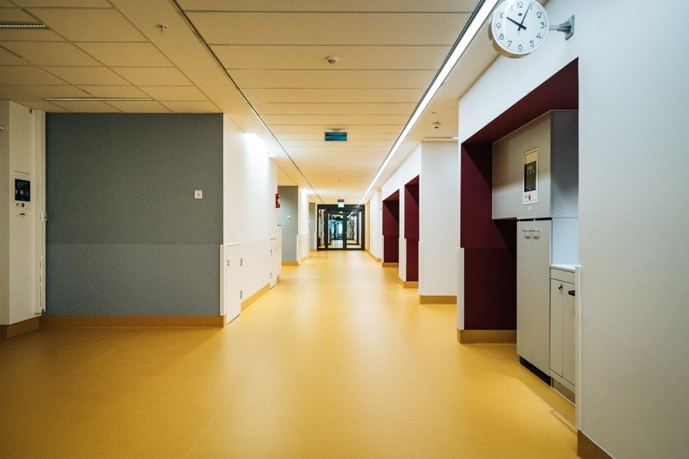 Modern hospital corridor with yellow flooring, white walls, and a clock showing 10:10.