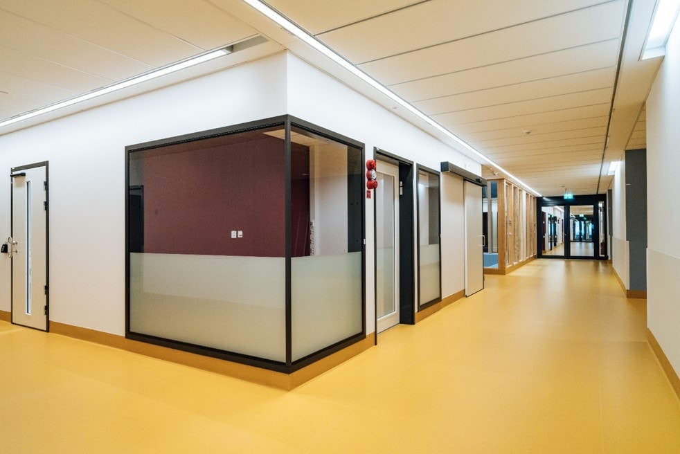 Modern hospital corridor with yellow flooring and glass-walled rooms.