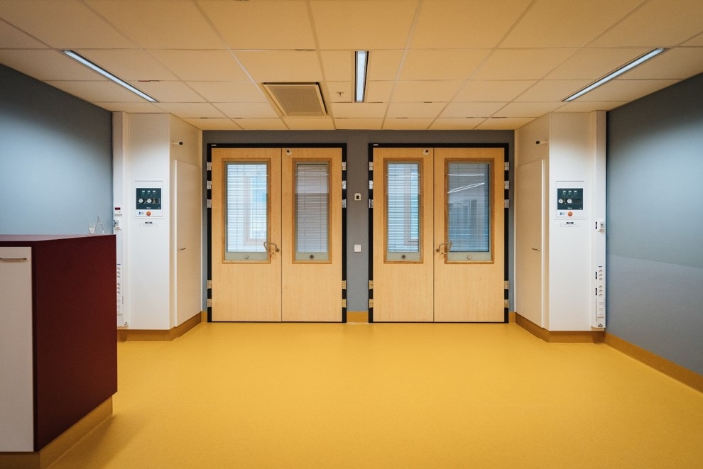Modern hospital corridor with double wooden doors, yellow flooring, and gray walls.