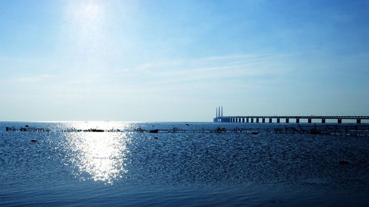 The Öresund Bridge Paisaje marino soleado con un puente distante bajo un cielo azul claro que se refleja en aguas tranquilas.