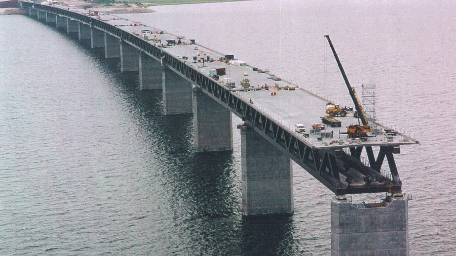 The Öresund Bridge Construcción de un largo puente sobre agua con grúas y vehículos en la plataforma sin terminar.