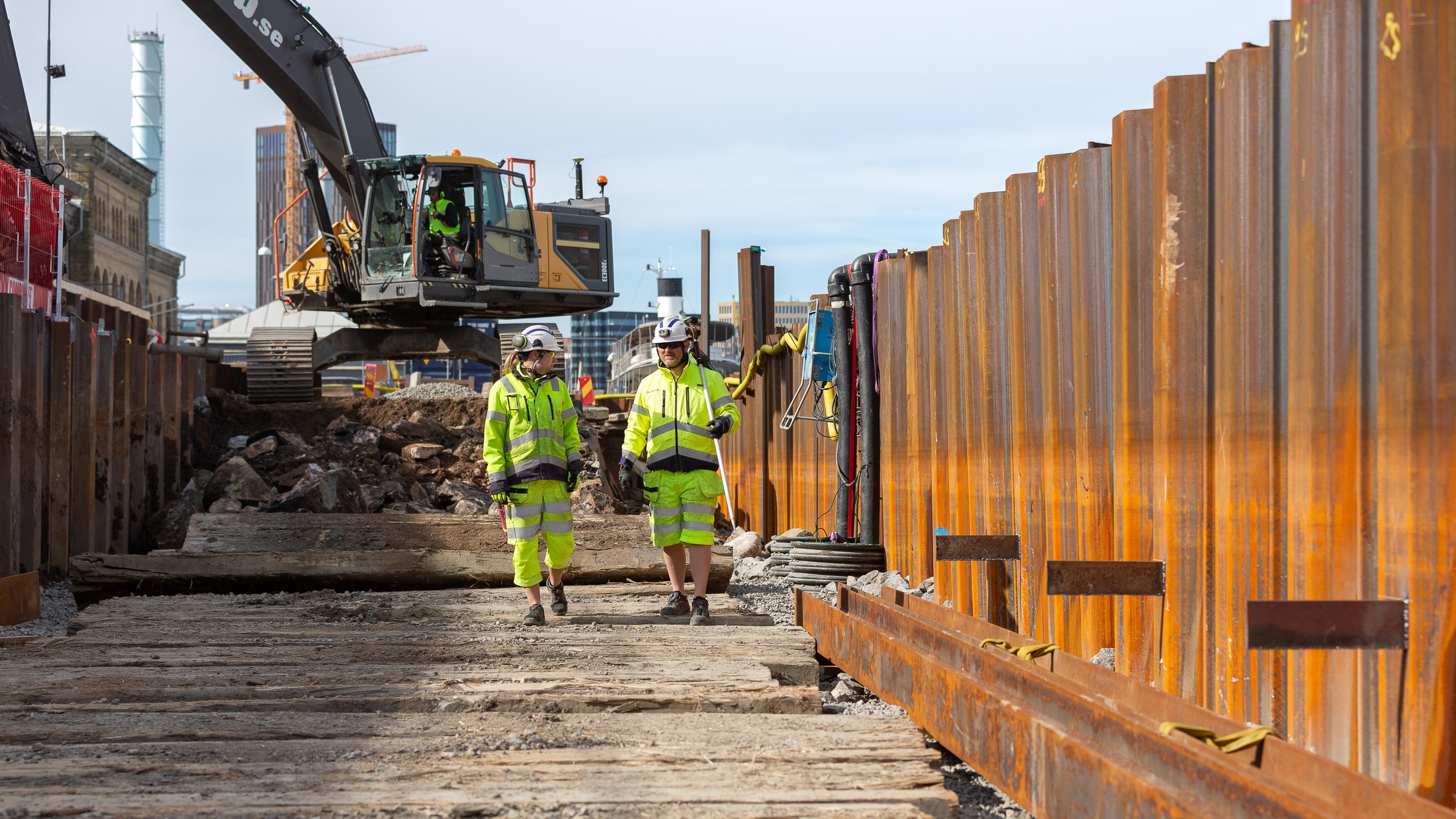 The project started in 2019. Two construction workers with an excavator in the background.