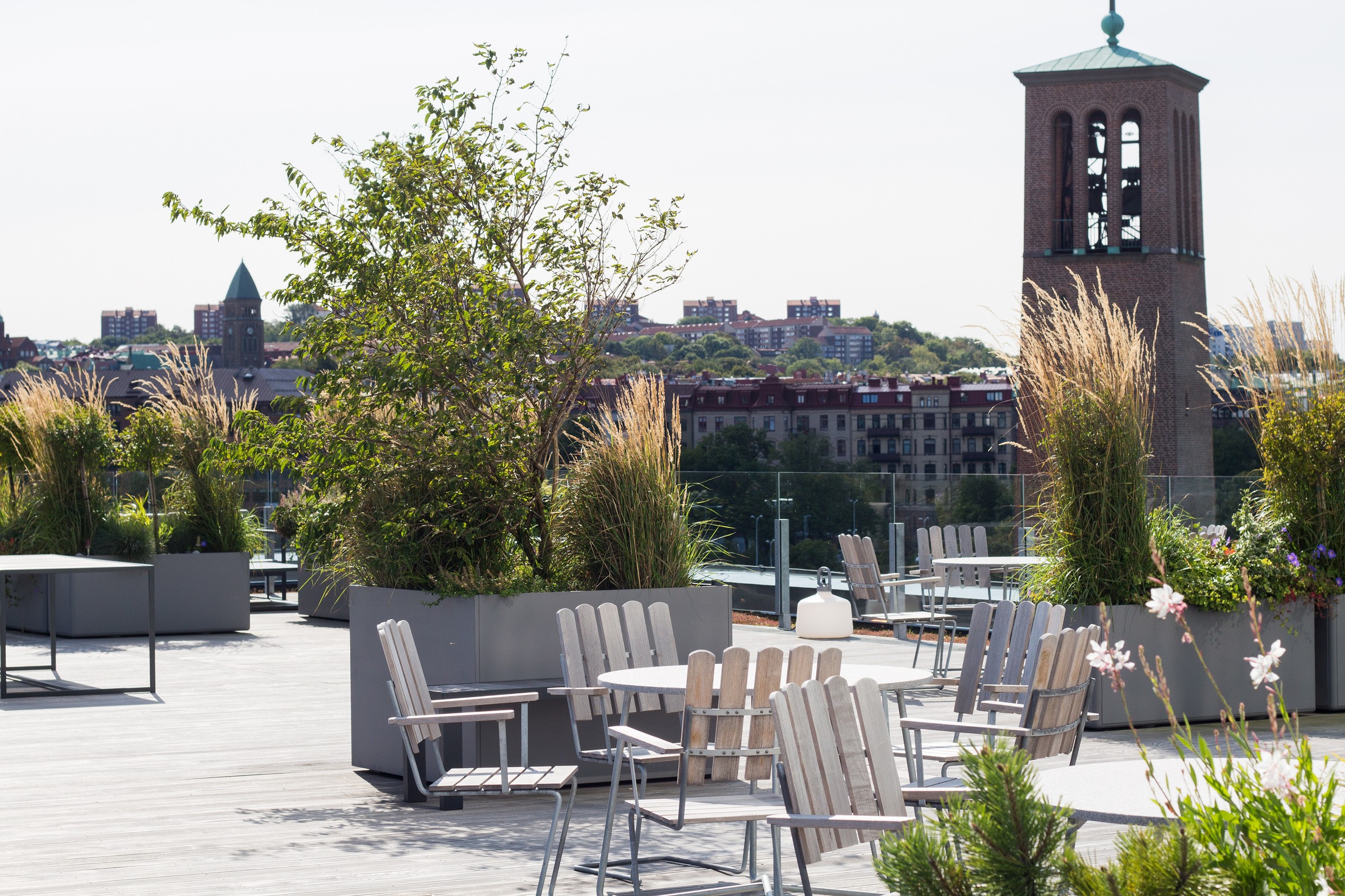 Rooftop terrace with modern seating and lush greenery, overlooking urban skyline and bell tower.