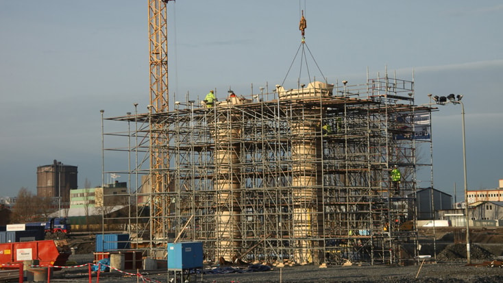 Partihallsförbindelsen Bridge Construction site with scaffolding and crane lifting materials, industrial buildings in the background.