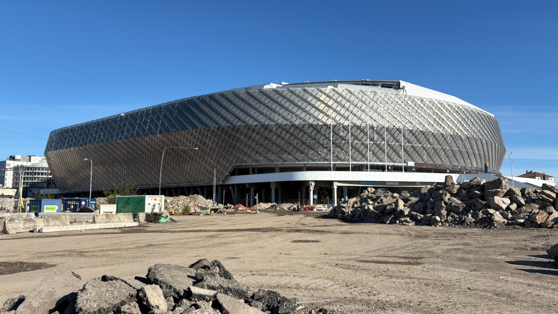 The Slakthusområdet in southern Stockholm is being transformed into a new district with electrified infrastructure. A large arena with a patterned facade is visible in the background, in front of a construction site with a gravel area, construction cabins, and large piles of stone and demolition material.