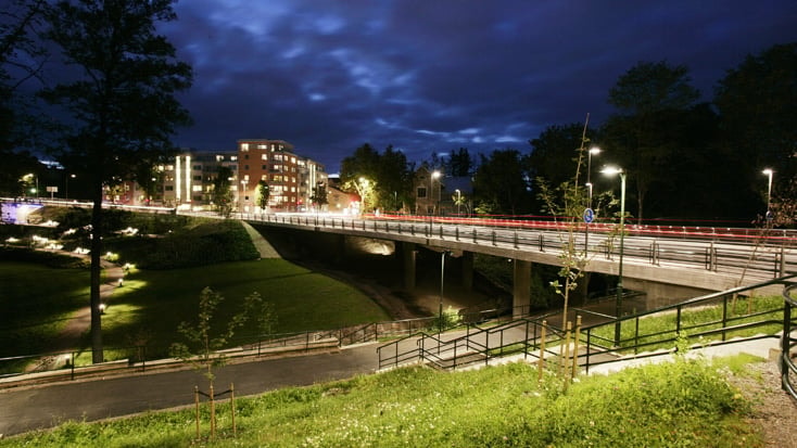 Night view of a brightly lit modern bridge with surrounding greenery and urban buildings.