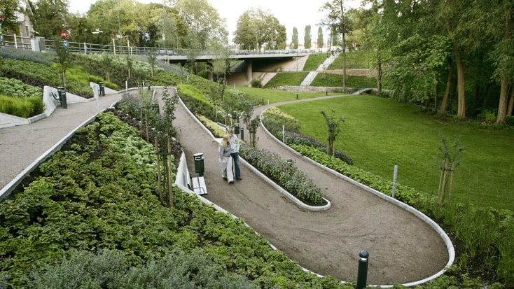 St Lars Bridge A scenic park pathway with lush greenery and a bridge in the background under a clear sky.