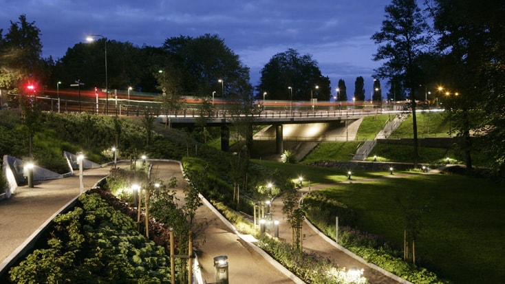 St Lars Bridge Illuminated park pathways and bridge at dusk surrounded by lush greenery and trees.