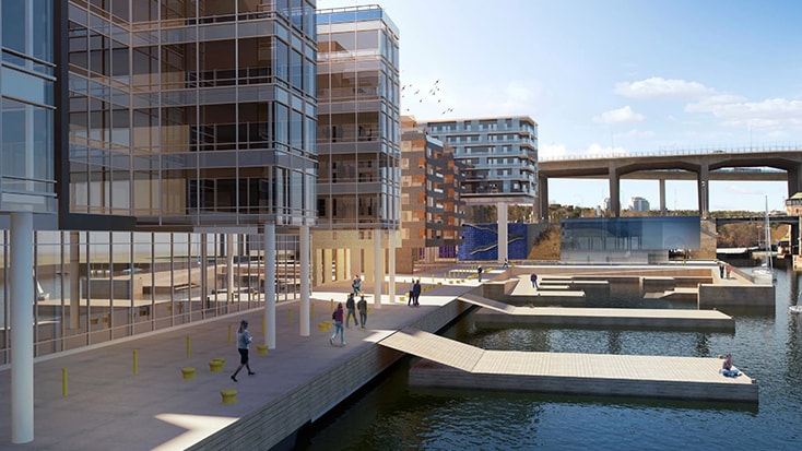The depot will not be visible from Hammarby Allé. Modern waterfront urban development featuring glass buildings, walkways, and a calm water area.