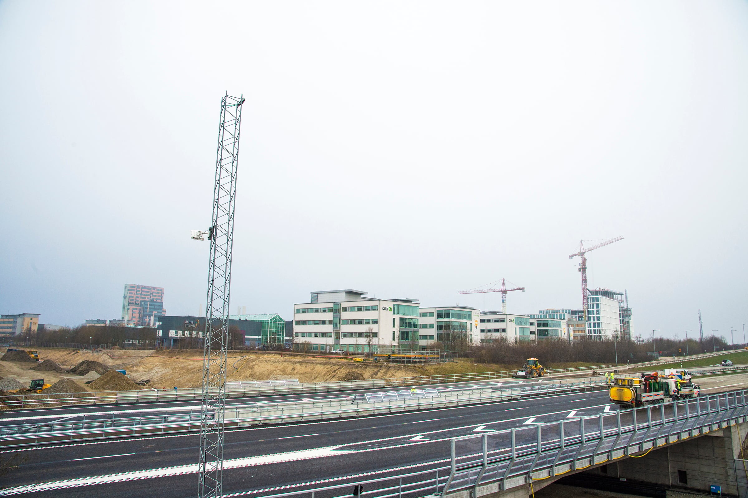 Modern highway construction with cranes and buildings under a cloudy sky.