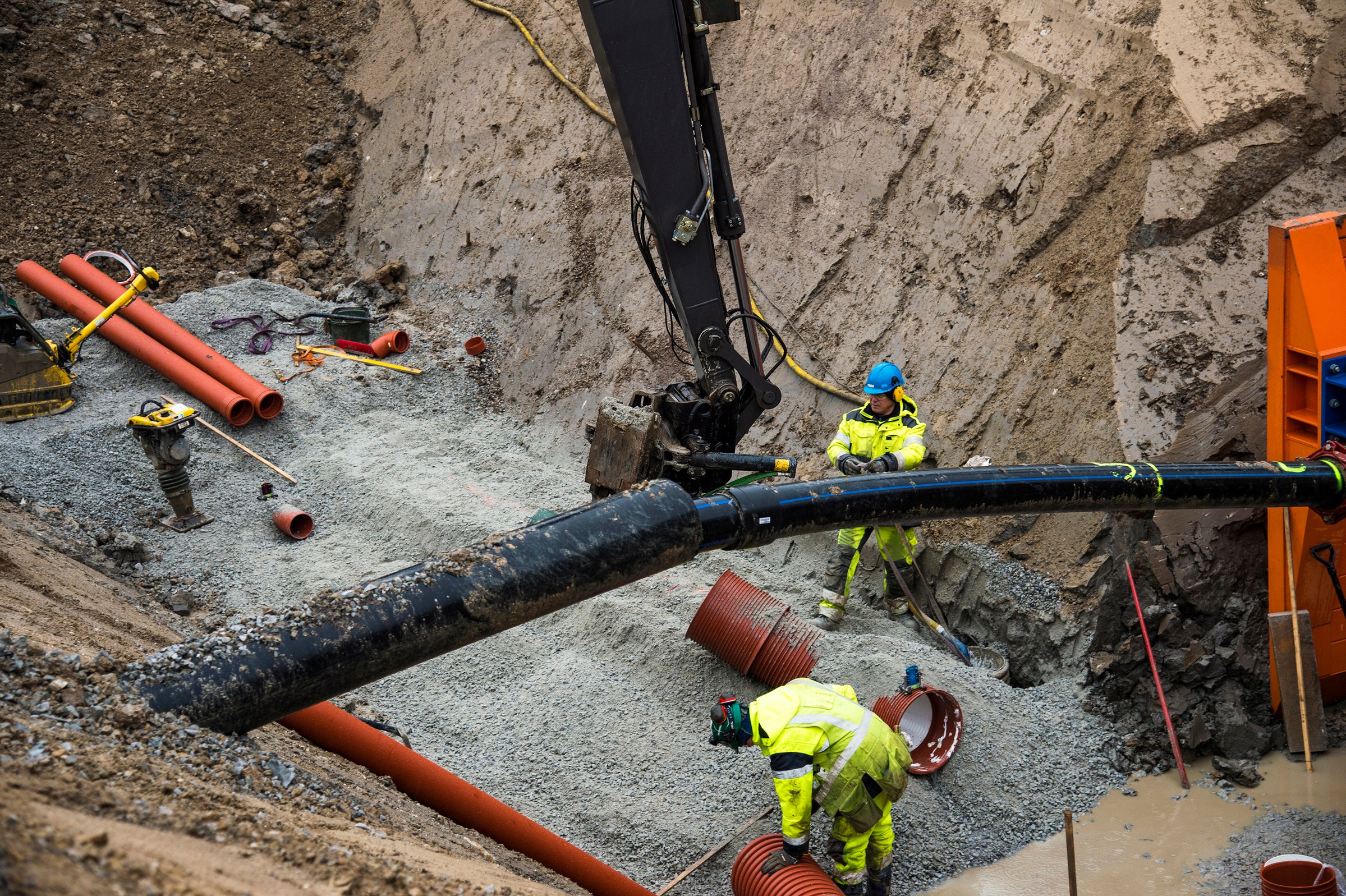 Construction workers in safety gear install underground pipes in a large excavation site.