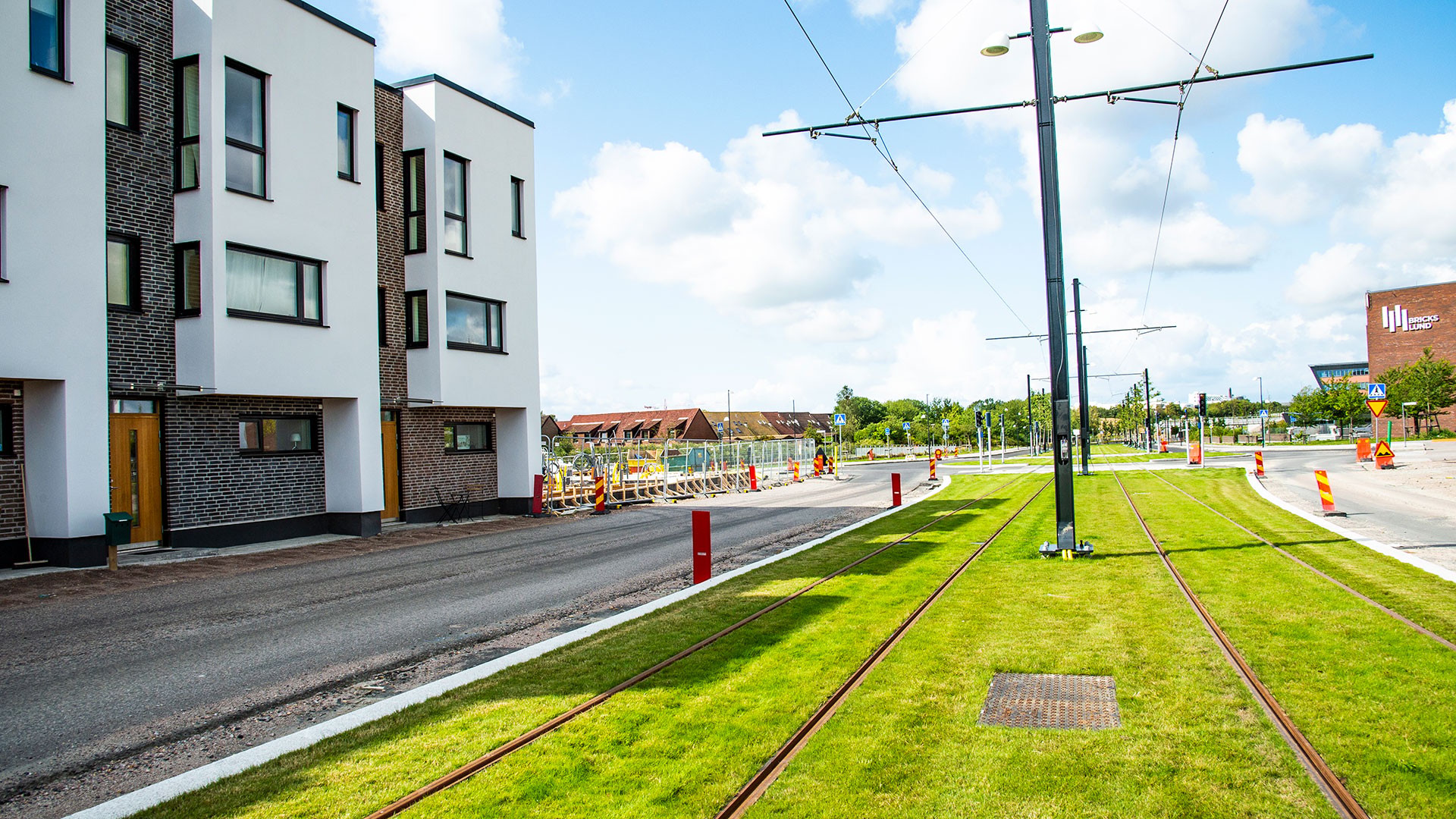 Modern row houses alongside a grassy tram track under a bright blue sky with scattered clouds.