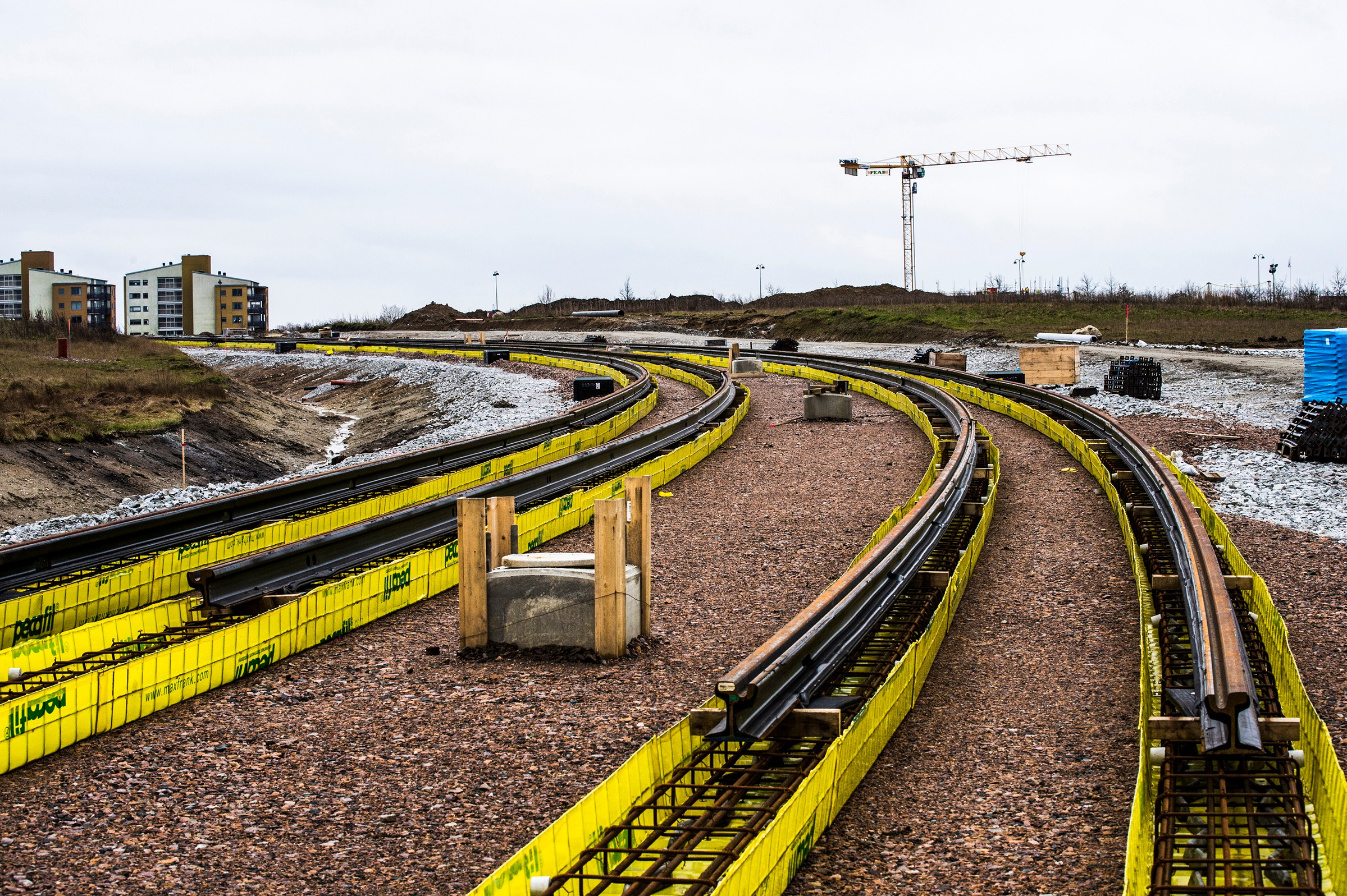 Construction site with curved railway tracks, surrounded by yellow barriers and distant crane.