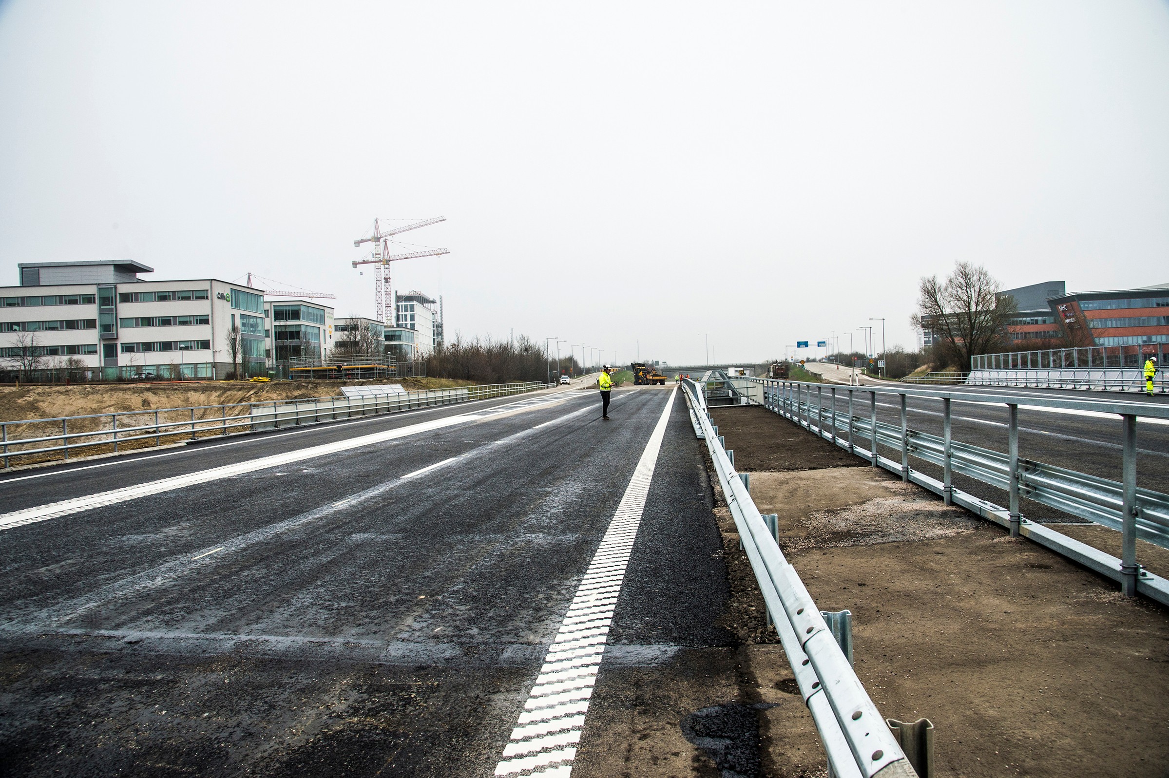 A newly paved highway with safety railings, surrounded by modern buildings and construction cranes.