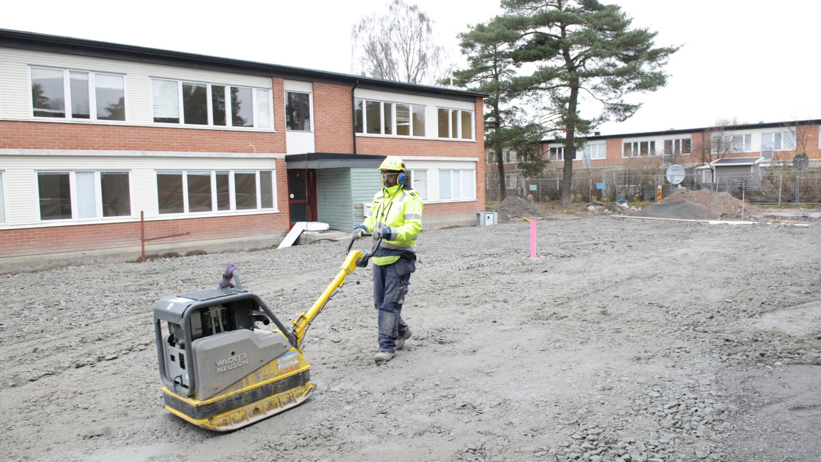 Construction worker operates a soil compactor on a gravel surface near a residential building.