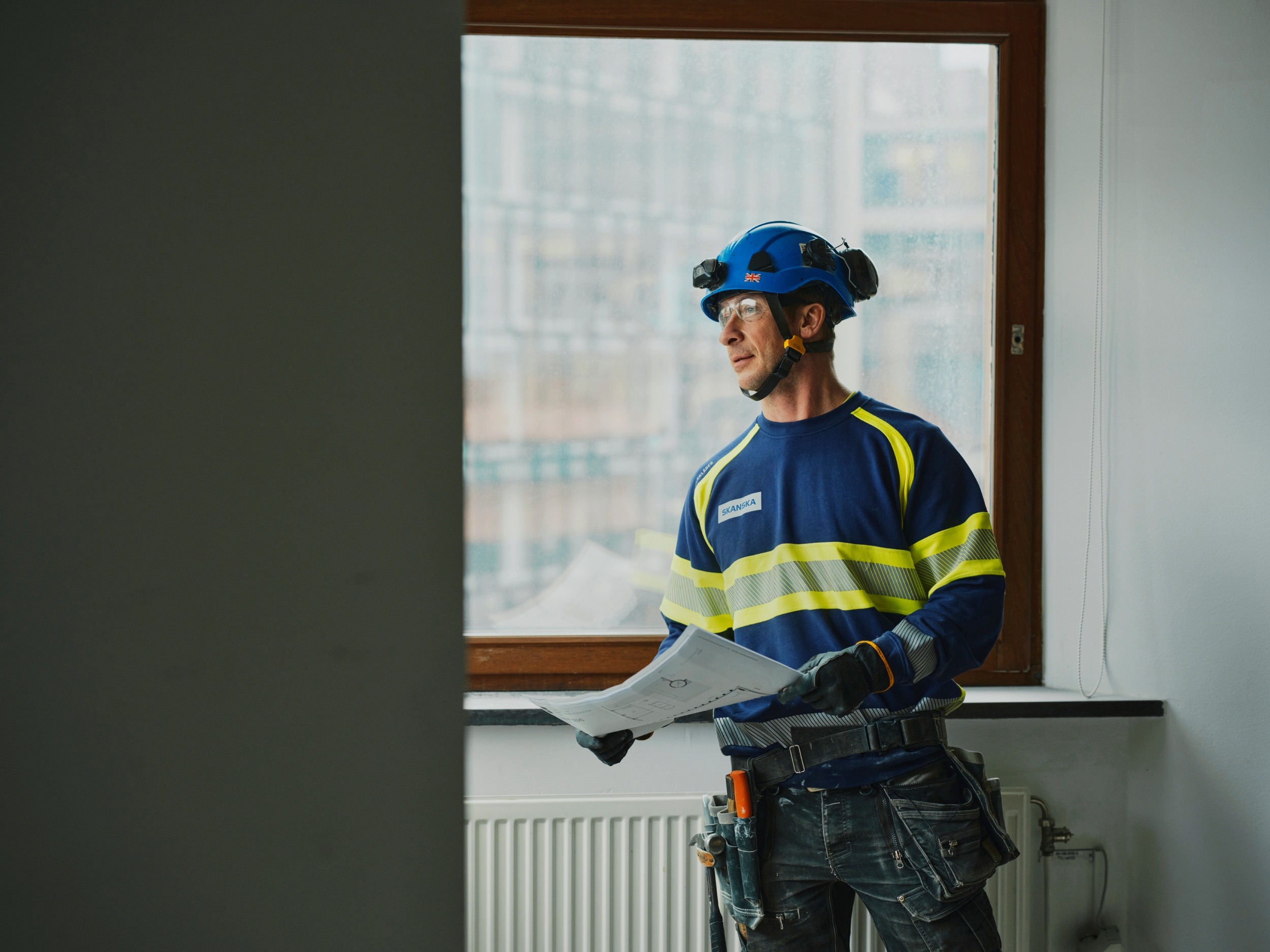 Construction worker in safety gear holding plans, standing near a window in a partially built room.