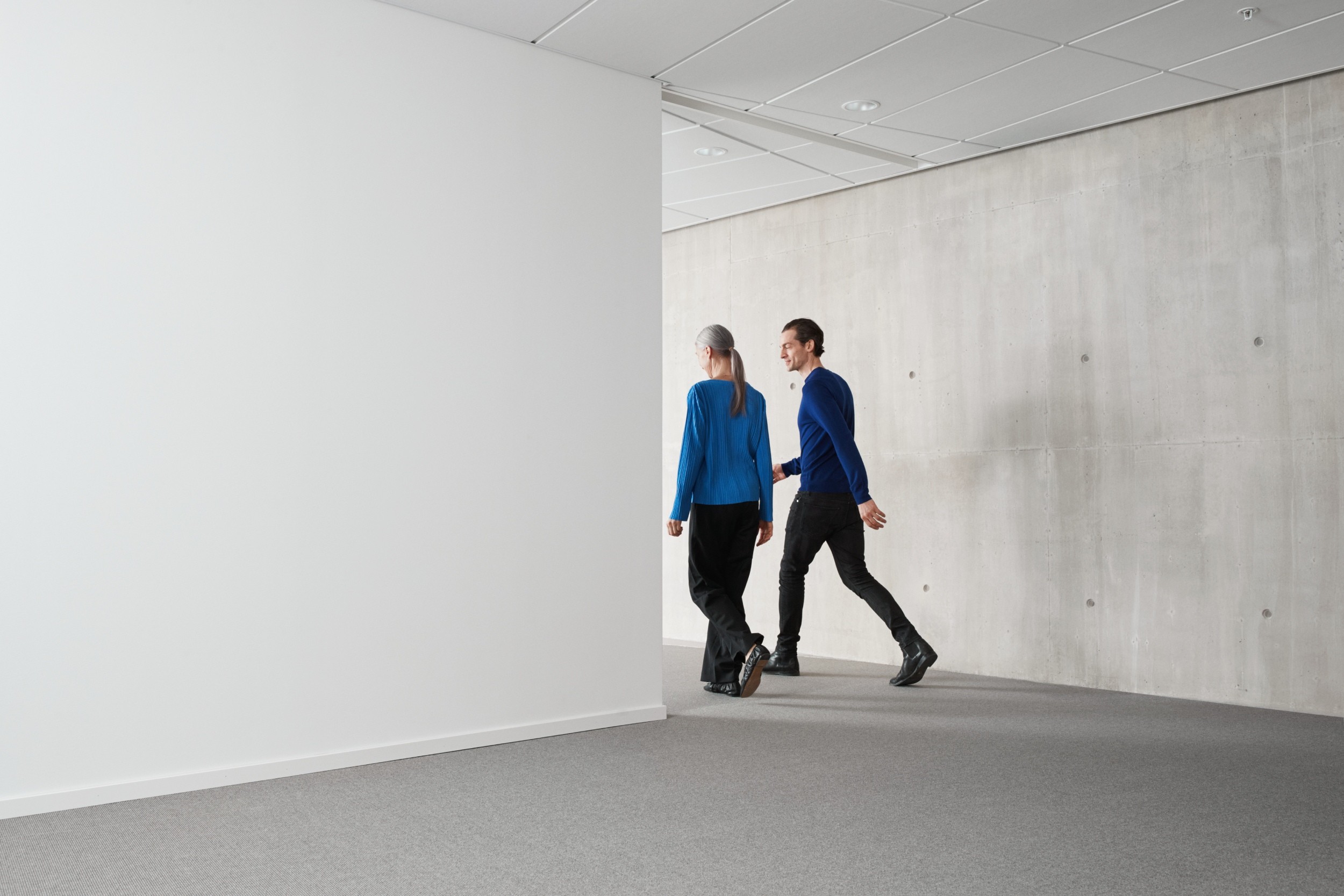 Two people in blue shirts walk down a modern hallway with gray carpet and concrete walls.