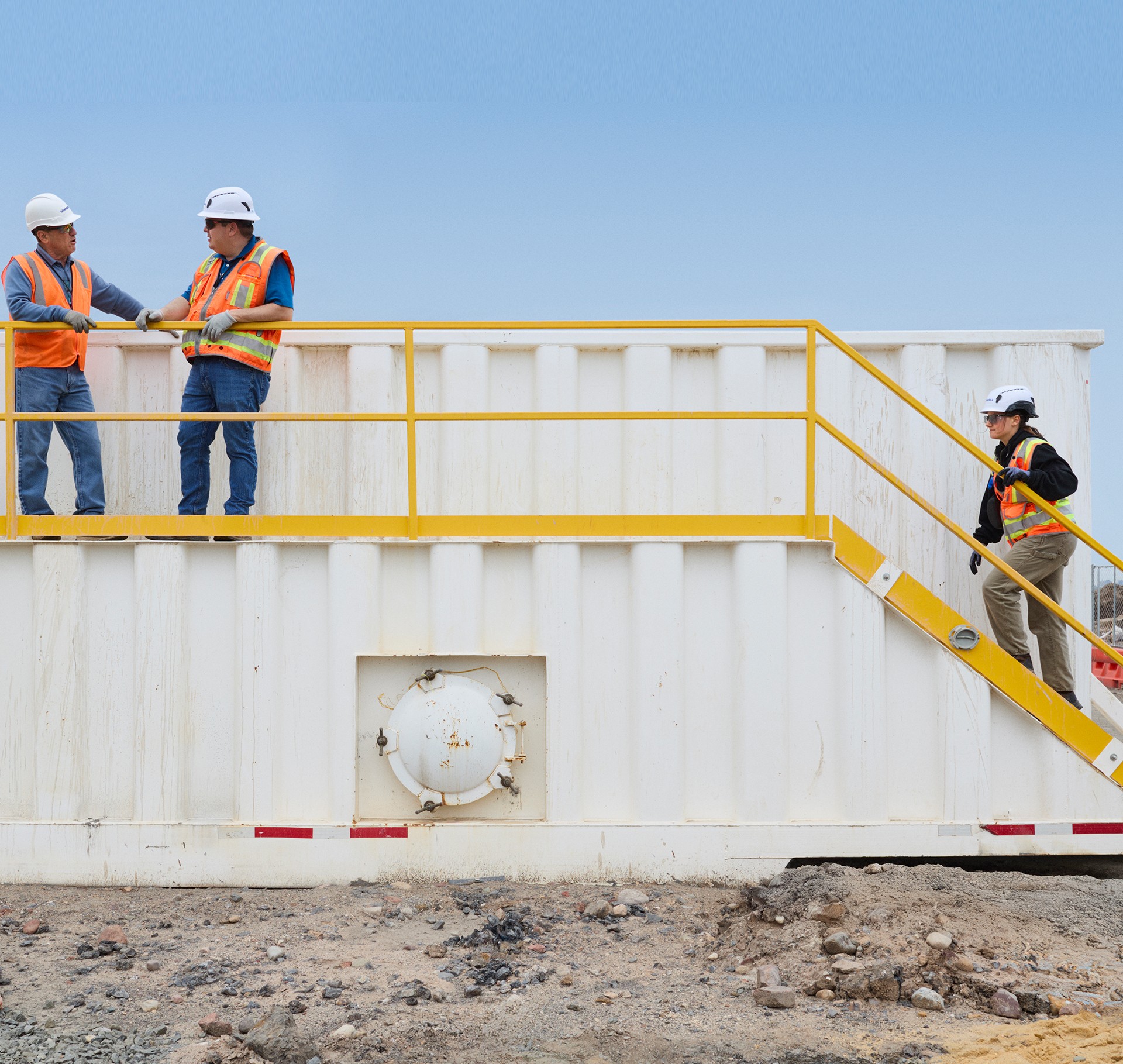 Construction workers wearing safety gear on industrial site stairs Two construction workers in safety gear on a yellow-railed platform against a clear sky.