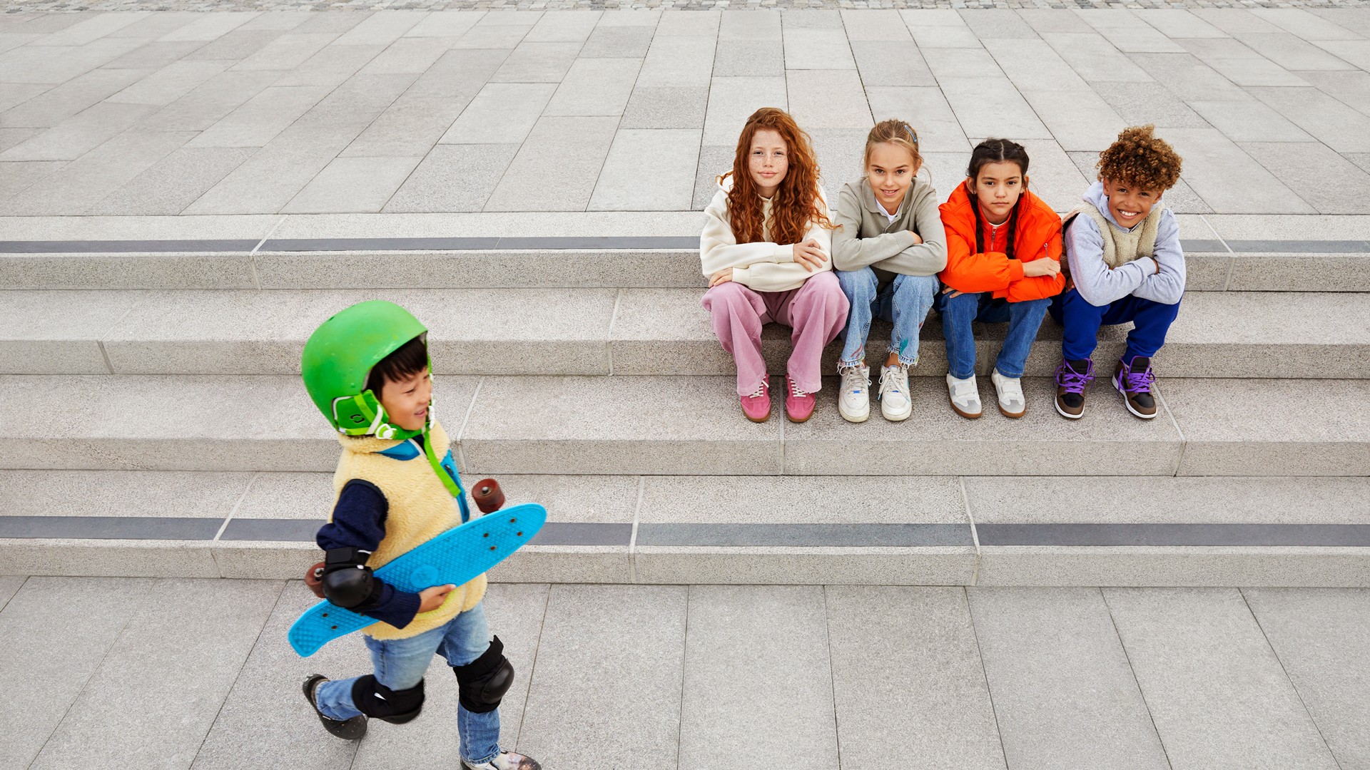 Child carrying skateboard approaches group of kids on steps A young child wearing a green helmet walks while holding a blue skateboard, approaching a group of kids seated together on wide concrete steps in an outdoor urban area.