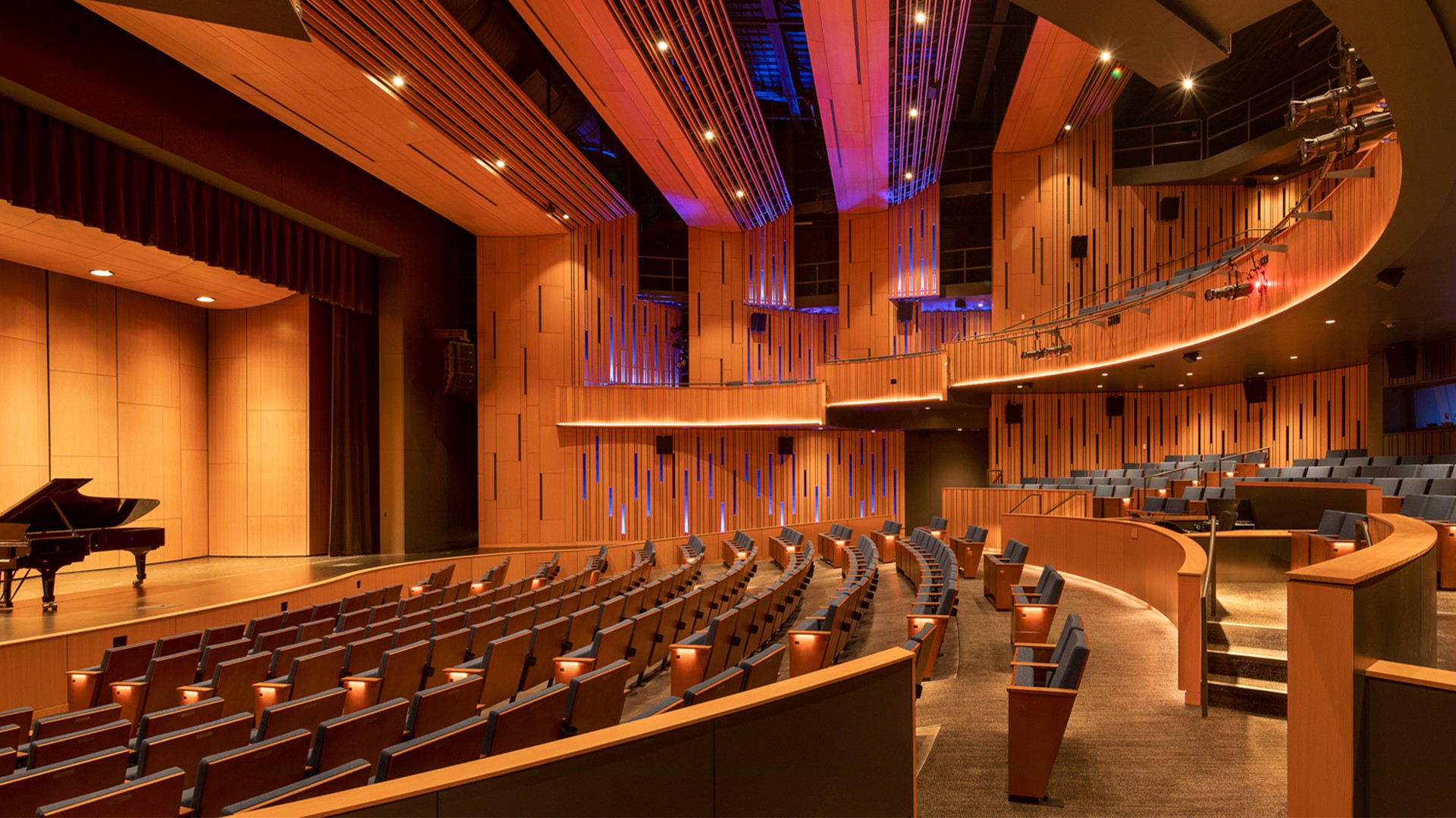 Modern theater interior with wooden paneling, curved seating, and a grand piano on stage.