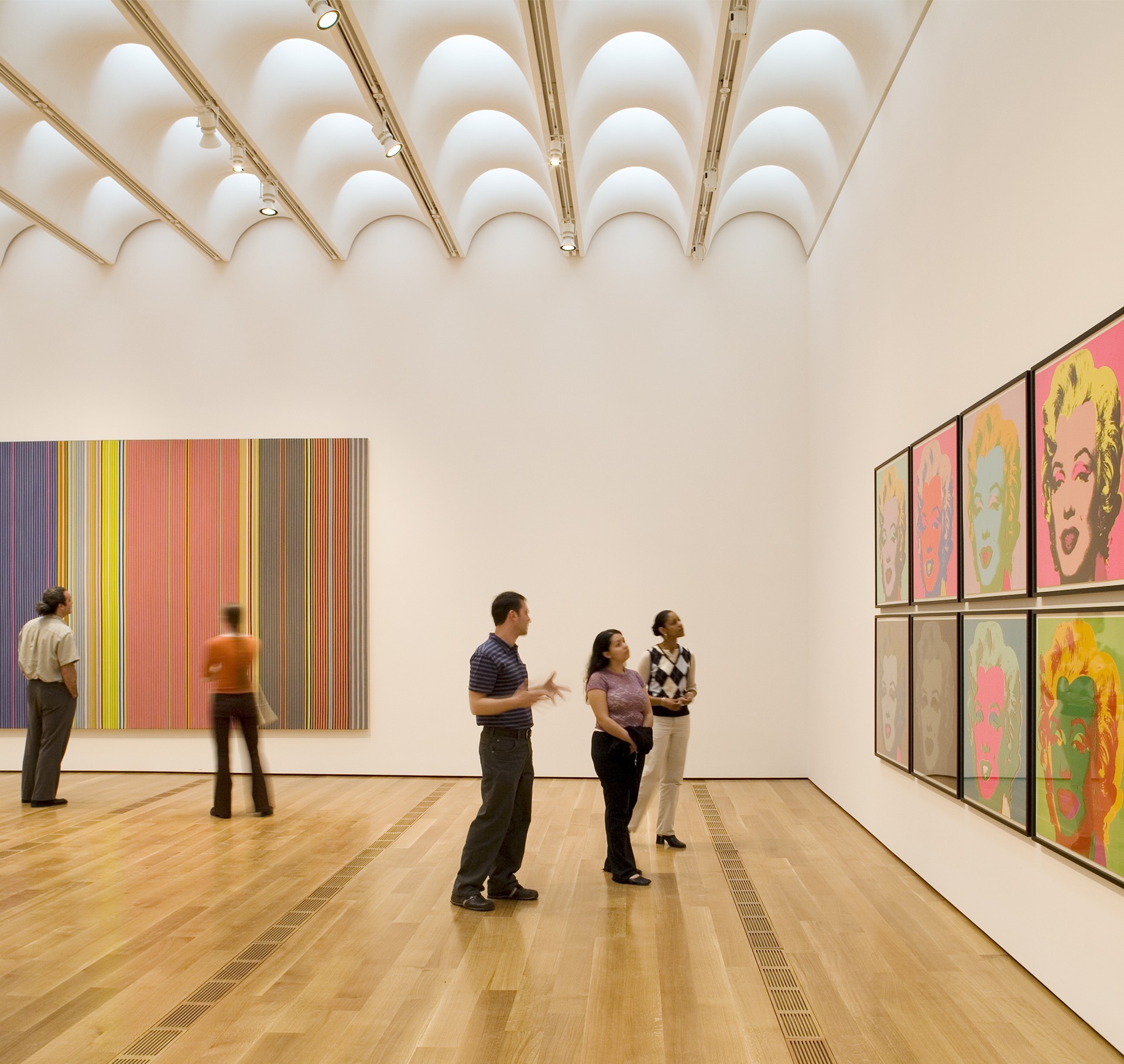 People admire colorful artwork in a modern gallery with unique ceiling design and wood flooring.