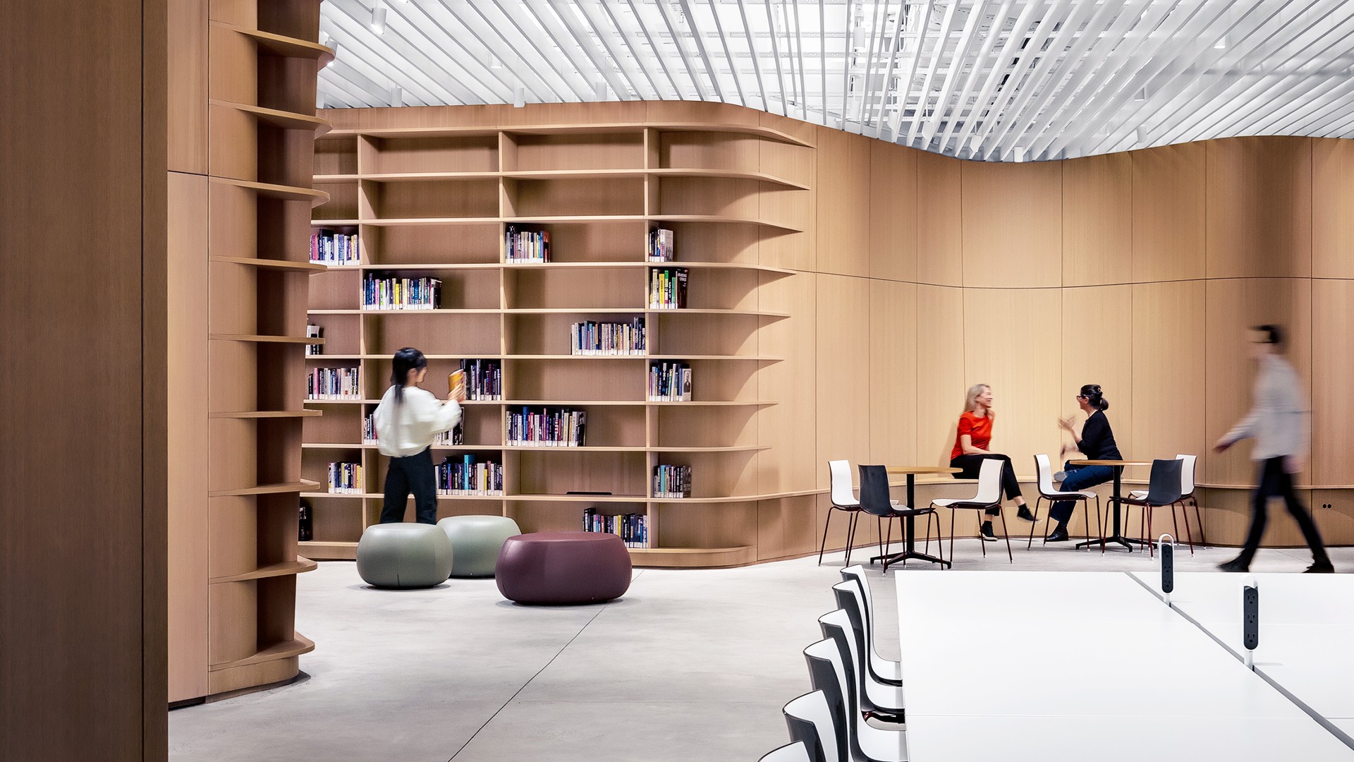 Modern library interior with wooden shelves, seating, and people working and walking.