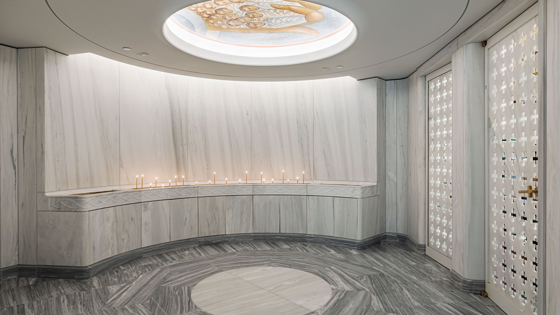 Interior of a modern marble chapel with lit candles and a circular skylight ceiling.