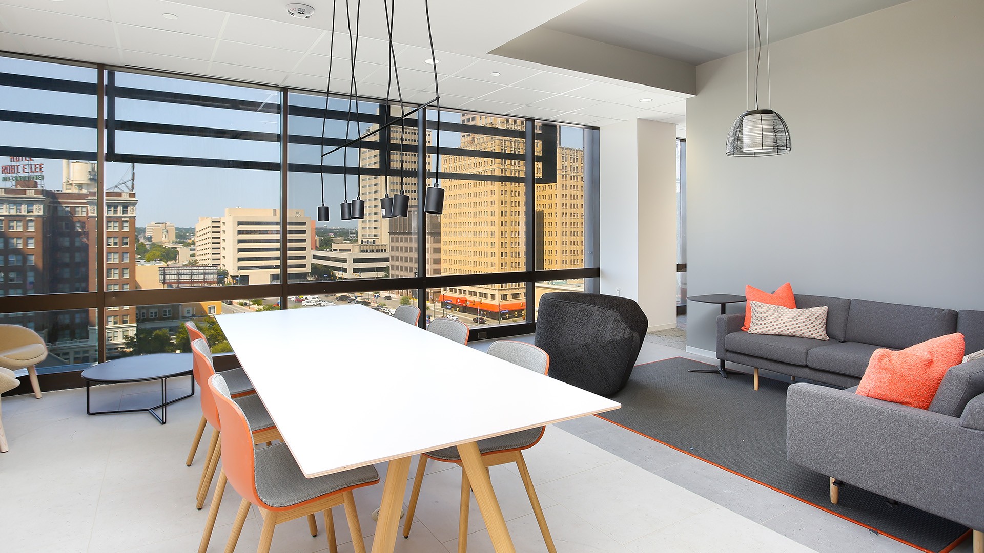 Modern office meeting room with large windows, white table, and gray sofas with orange pillows.
