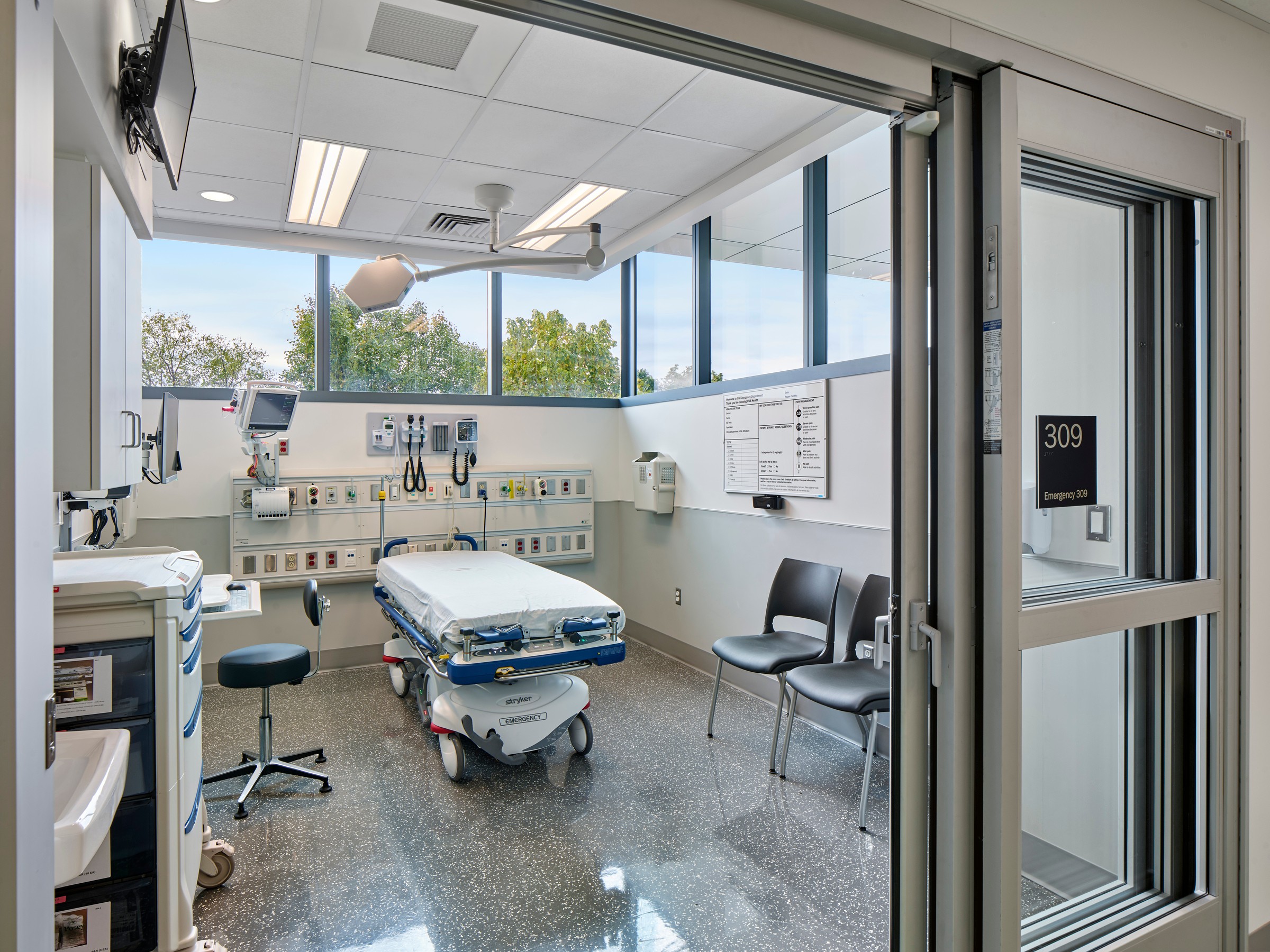 Modern hospital room with medical equipment, patient bed, chairs, and large windows.