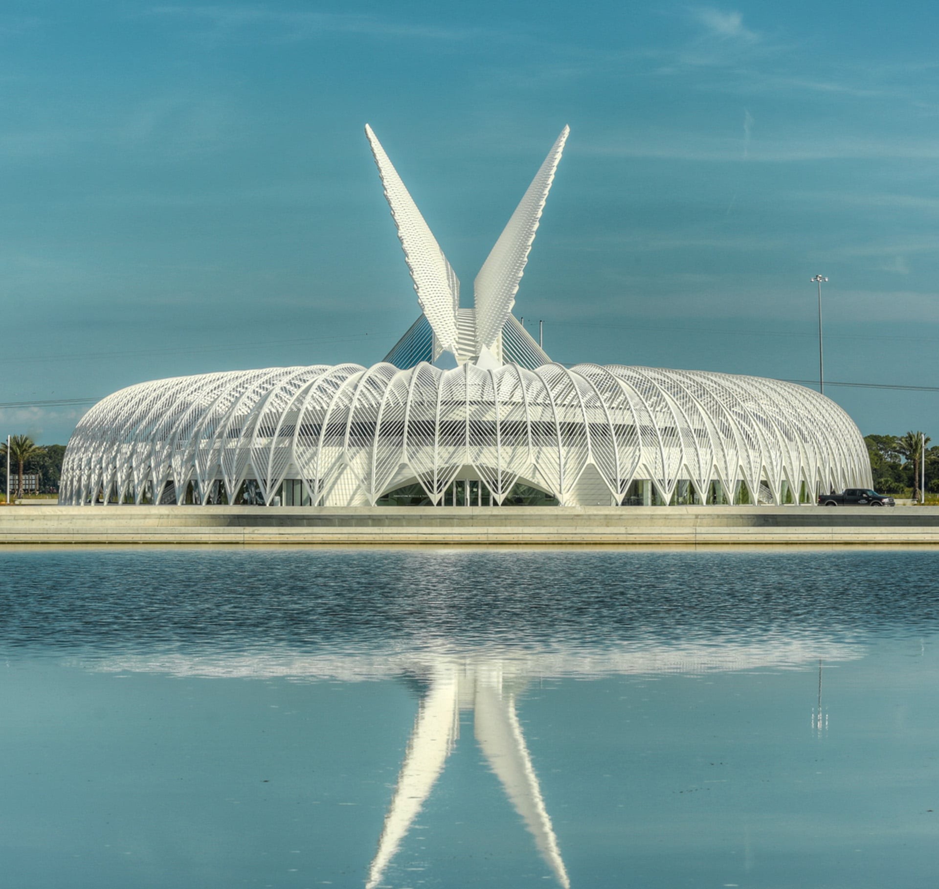 Futuristic White Architectural Pavilion with Water Reflection Futuristic white building with wing-like structures, reflected in a serene lake under a blue sky.