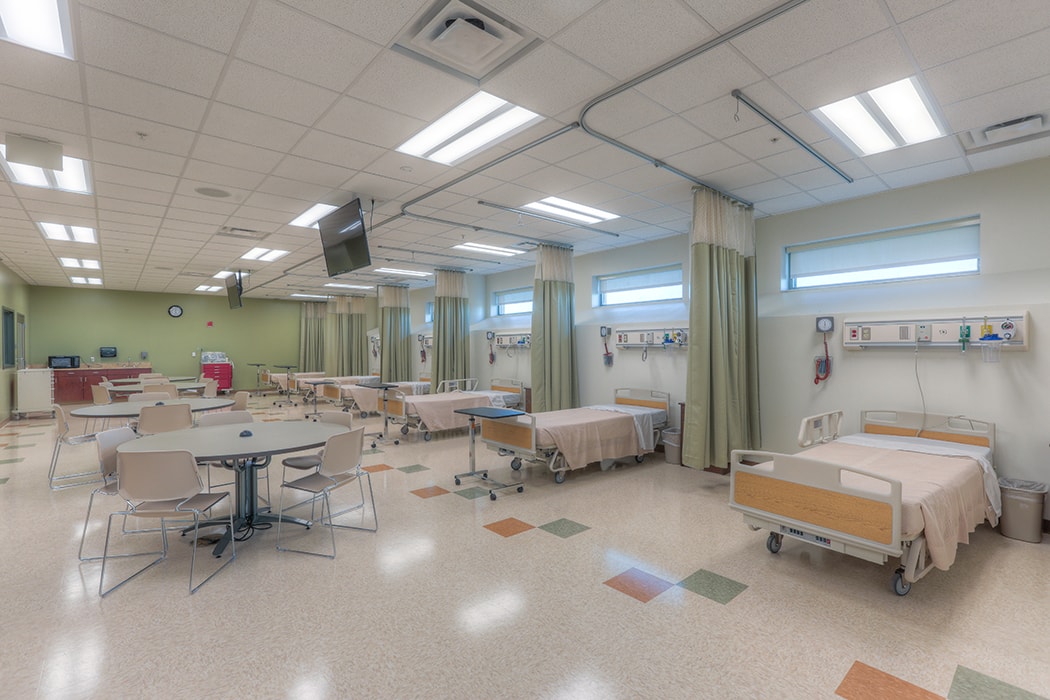 Hospital ward with multiple beds, curtains for privacy, tables, and bright overhead lighting.