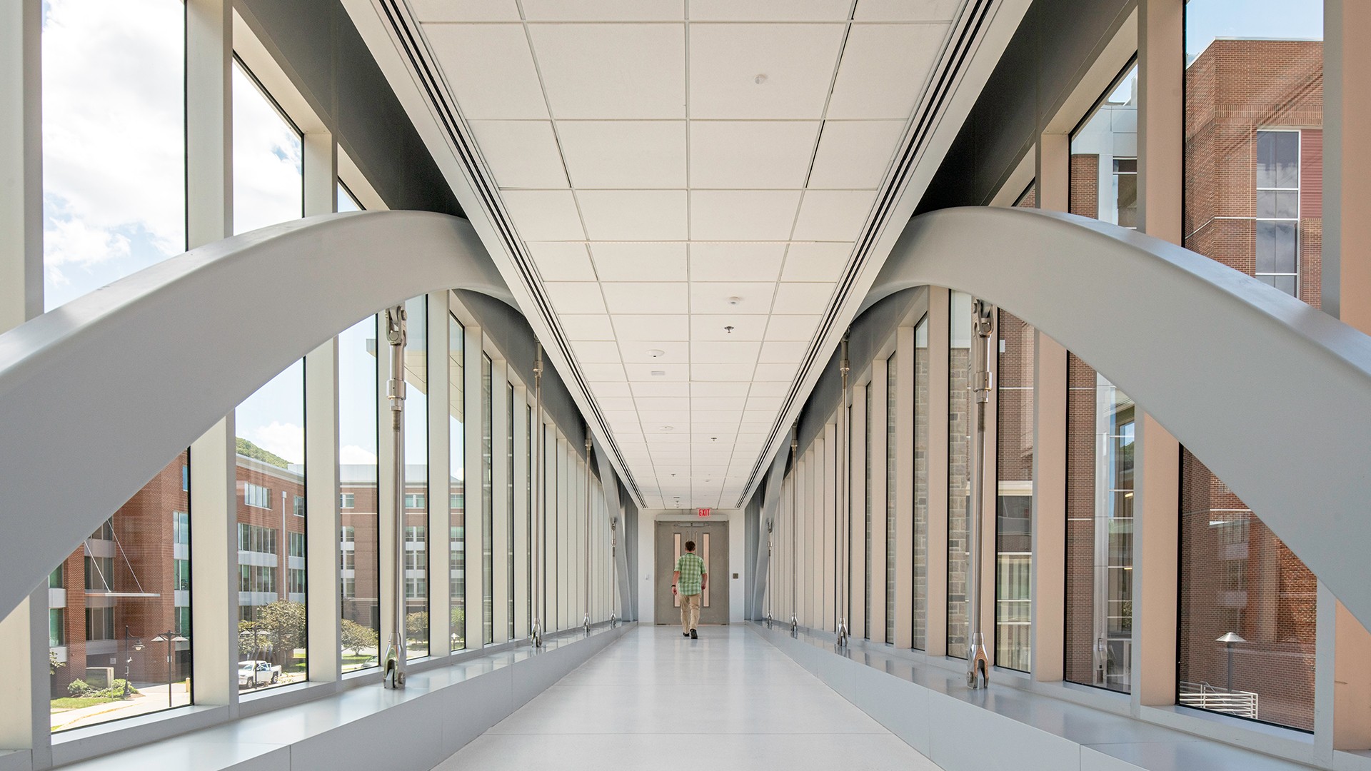 Modern glass corridor with a person walking, surrounded by office buildings outside.