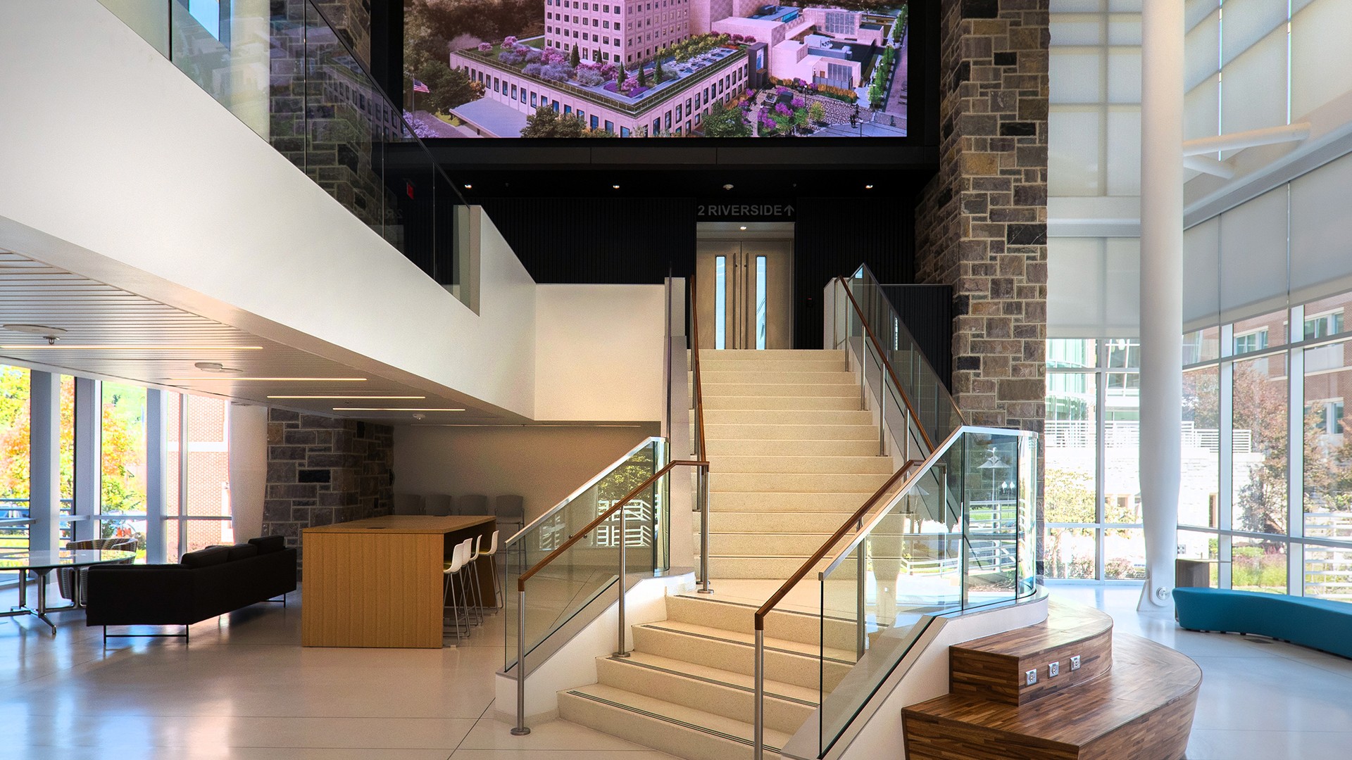 Modern office lobby with staircase, glass railings, and large digital display screen.