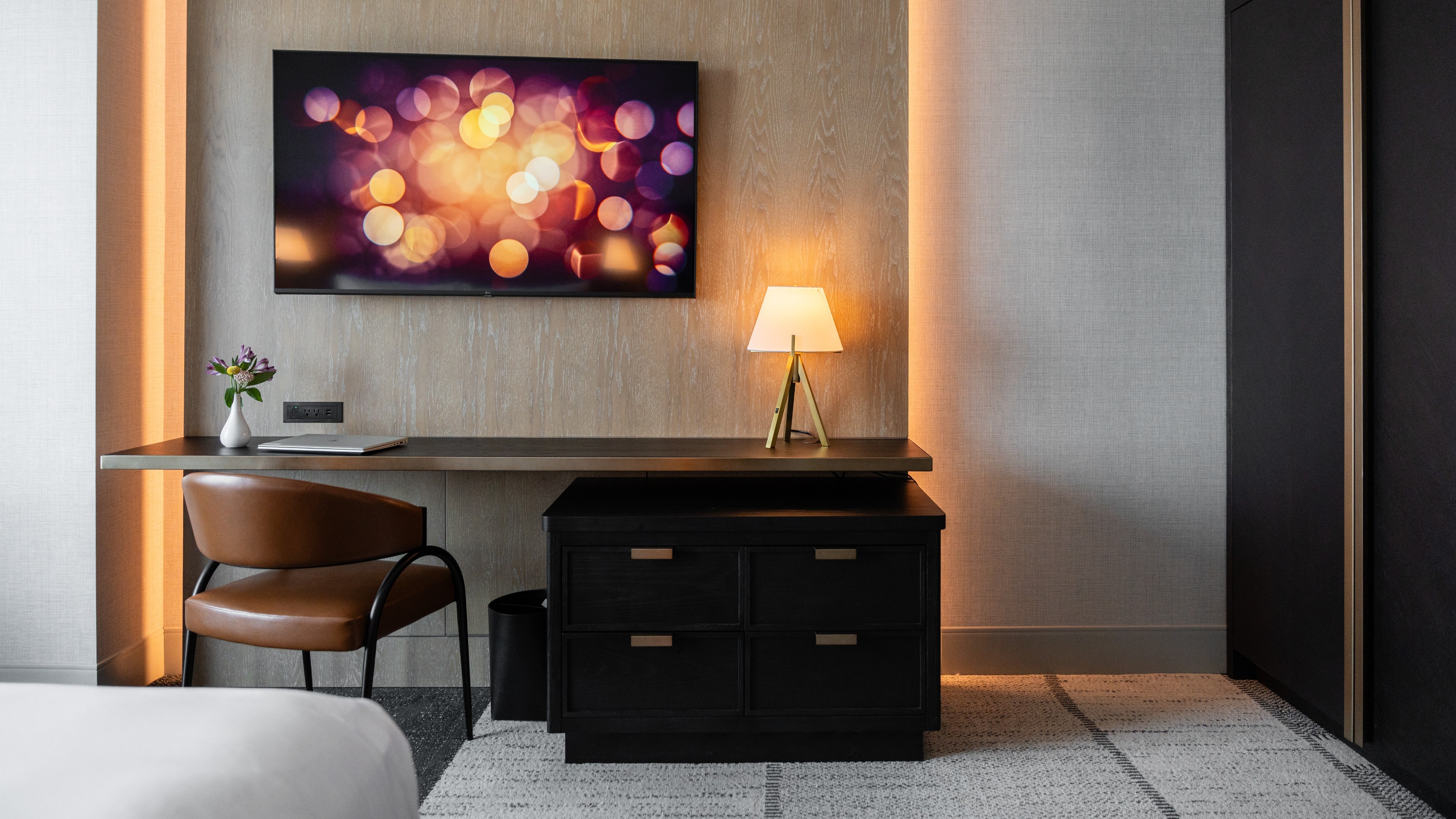 Modern hotel room workspace with a desk, leather chair, lamp, and wall-mounted TV.