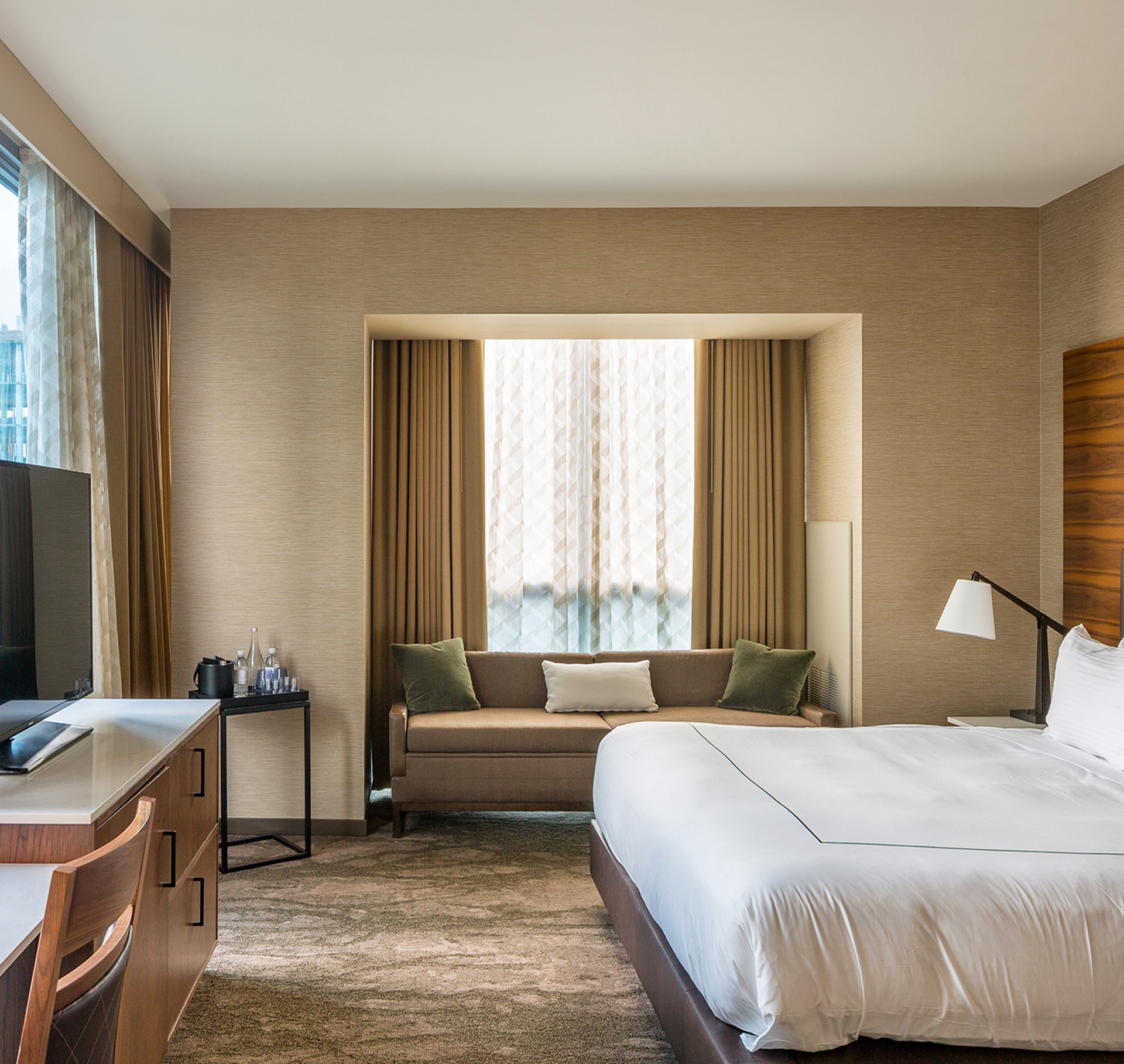 Modern hotel room with a large bed, sofa, desk, and TV, featuring warm, neutral tones.