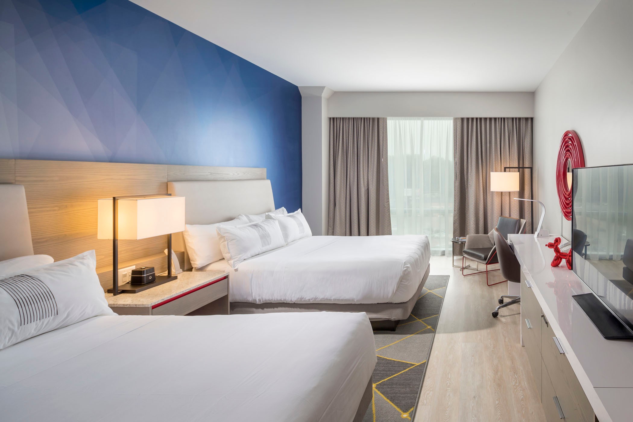 Moderne hotelkamer met twee tweepersoonsbedden, blauwe accentmuur en groot raam met gordijnen.