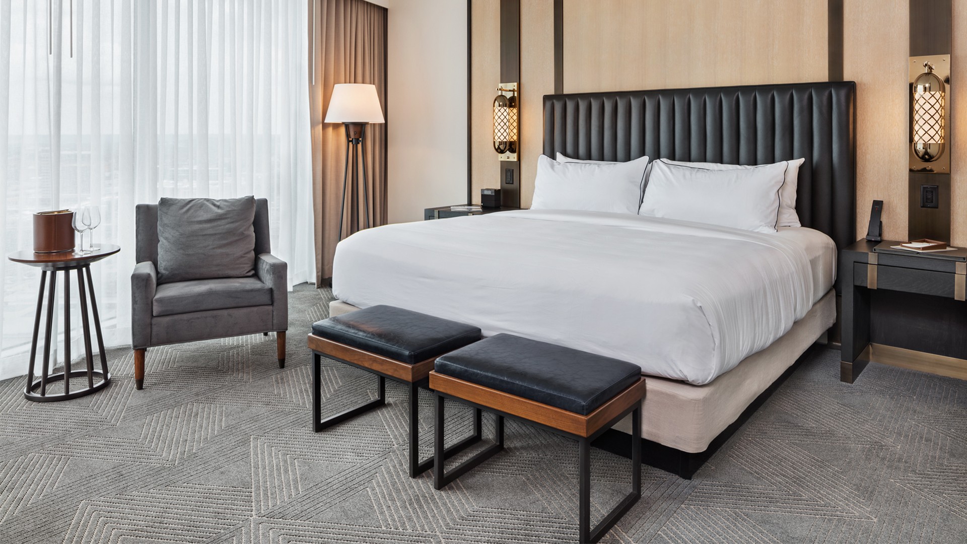 Modern hotel room with king-size bed, armchair, side tables, and soft lighting.