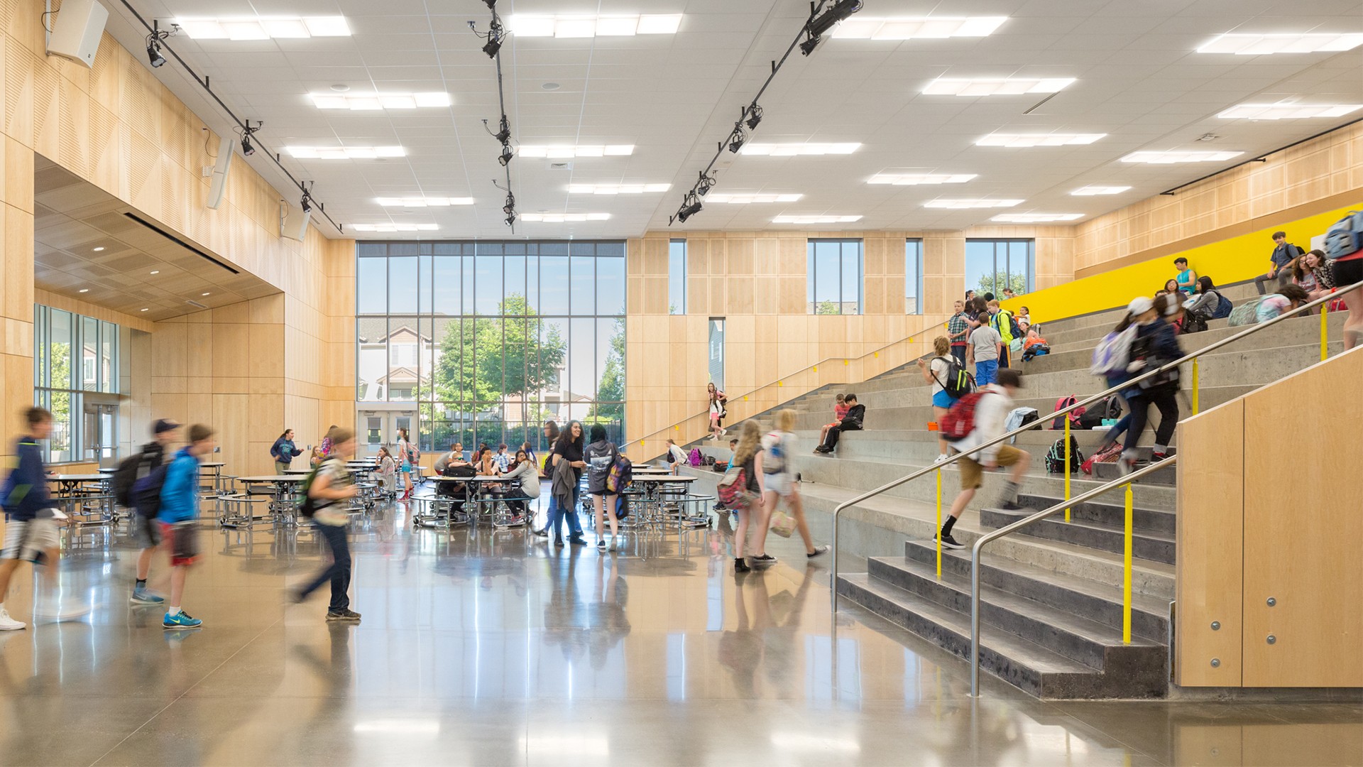 Students gather and walk inside a bright, modern school cafeteria with large windows.