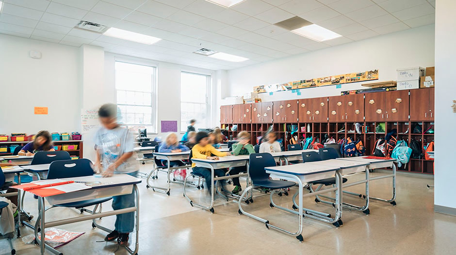 A bright classroom with children seated at tables, engaged in activities, and storage cubbies visible.