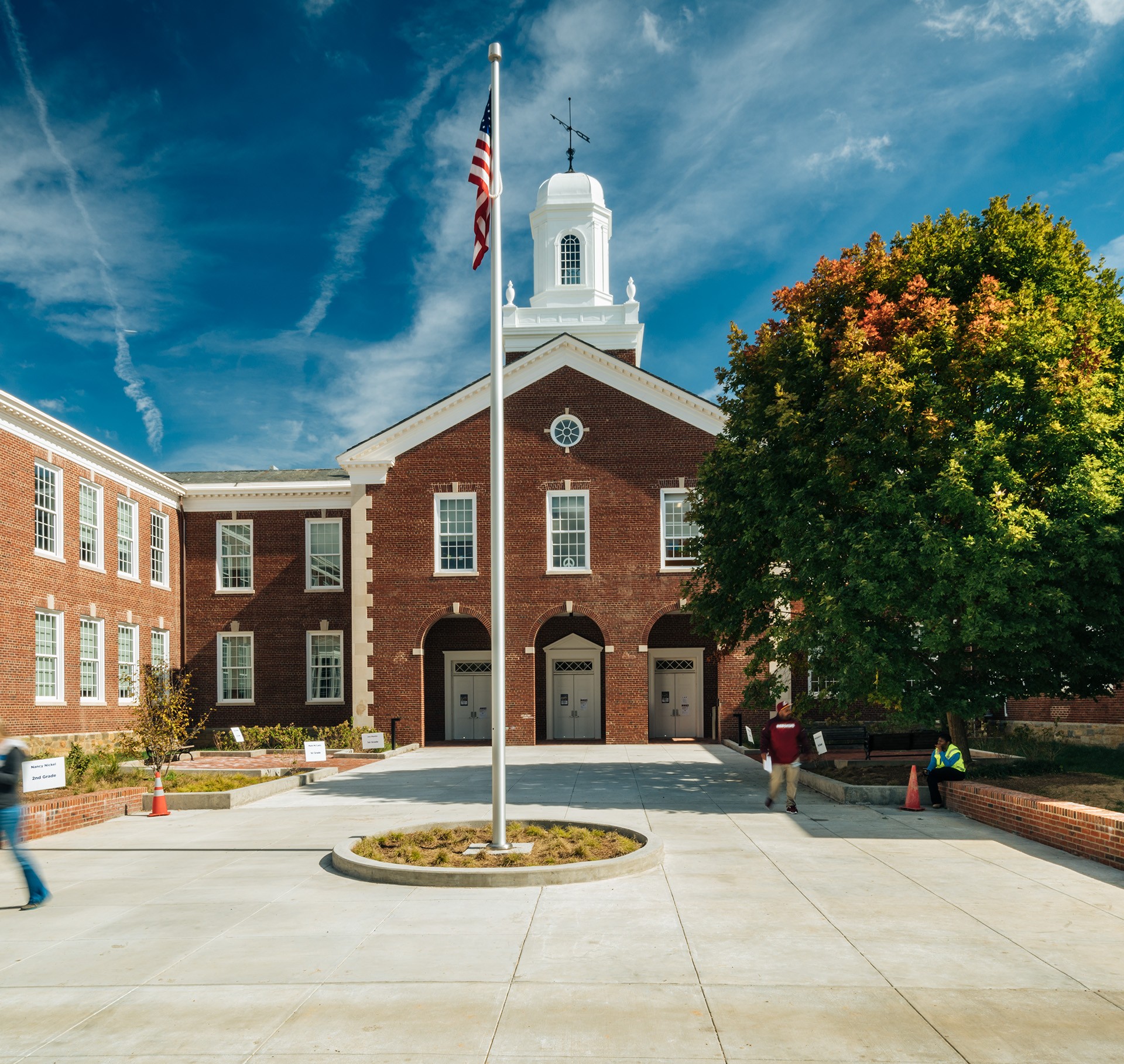Brick school building with steps, flagpole, and bicycles; people walking nearby under a blue sky.