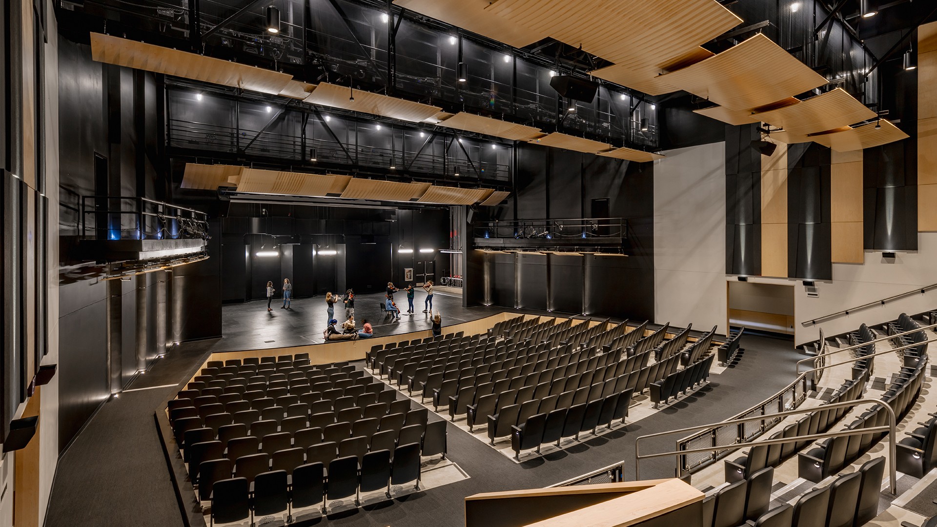 Modern theater auditorium with black seats, wooden ceiling panels, and a large stage.