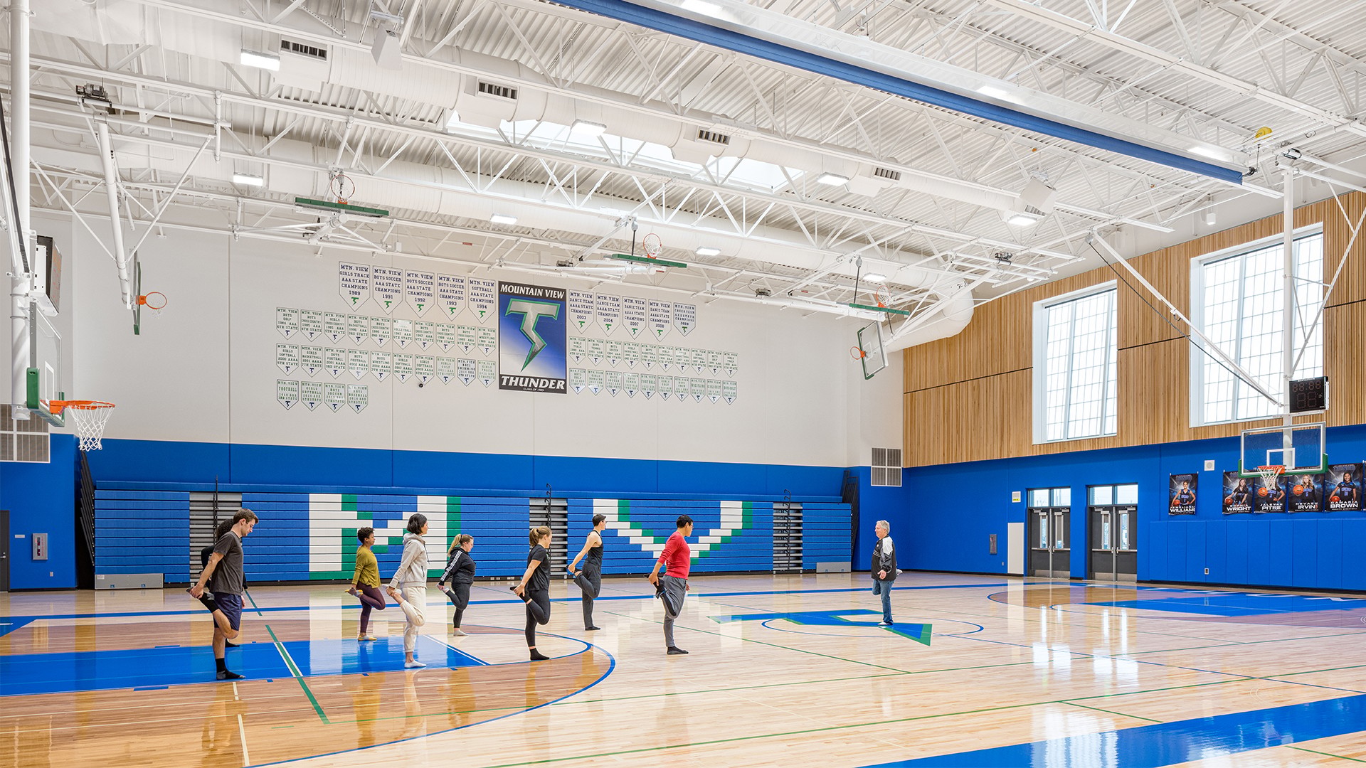 A group of people exercising with brooms in a bright, spacious school gymnasium.
