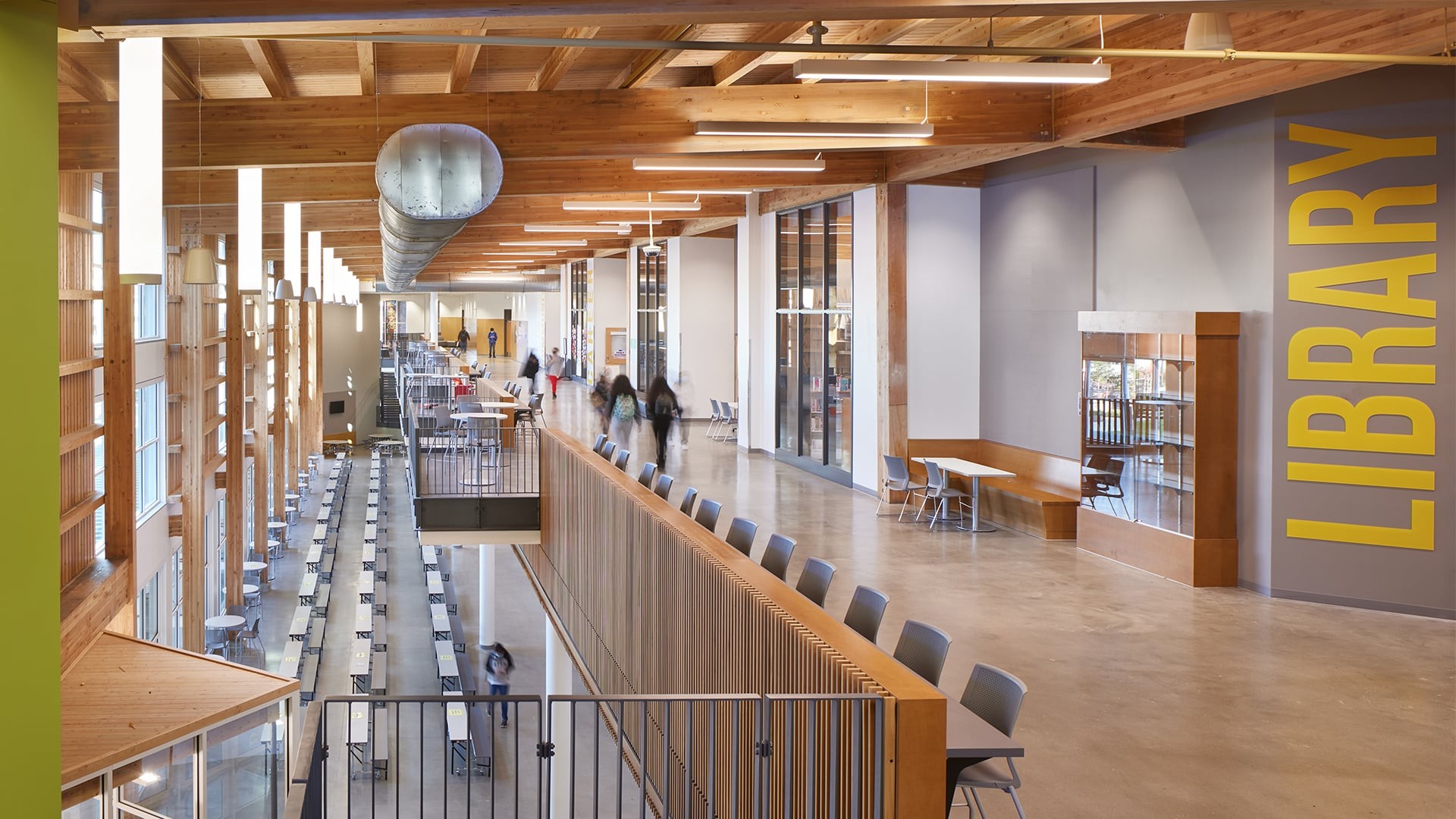Modern library interior with wooden beams, long tables, and people walking.